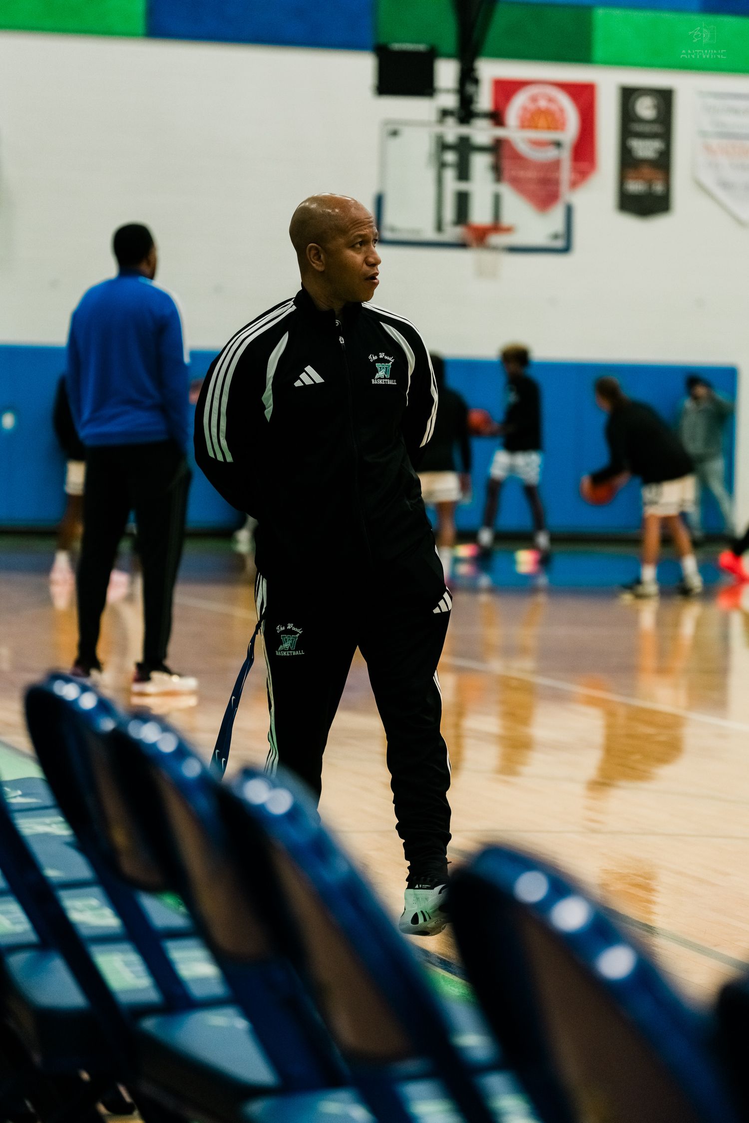 A man in a black Adidas tracksuit observes basketball warm-ups from the sideline of a gymnasium.