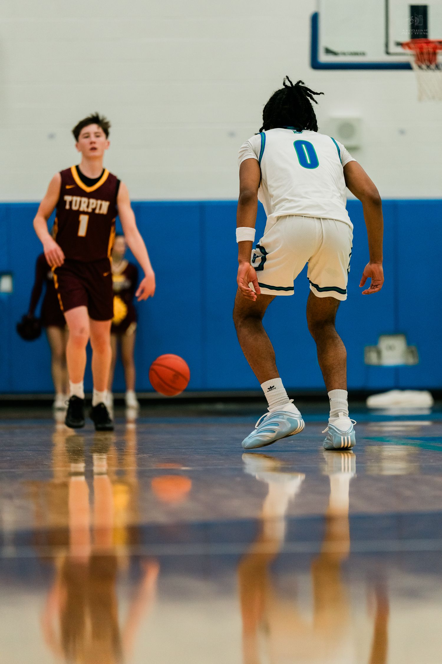 Player in white number 0 jersey dribbles toward Turpin player number 1 on reflective indoor basketball court.