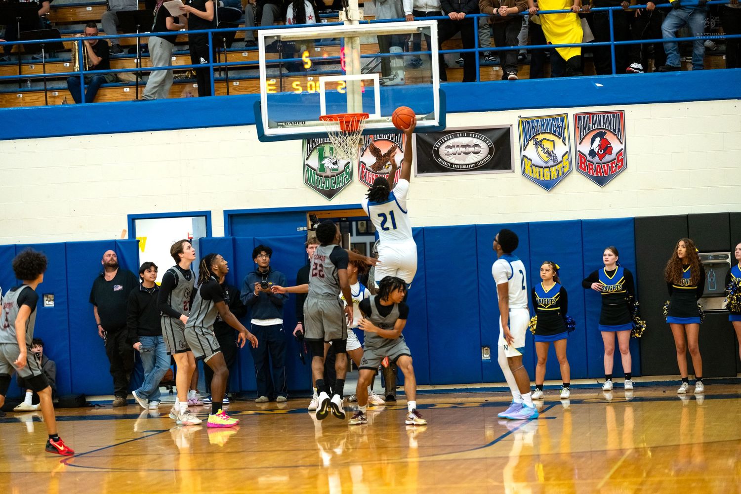 Basketball player number 21 in blue dunks over defenders during exciting high school gymnasium game.