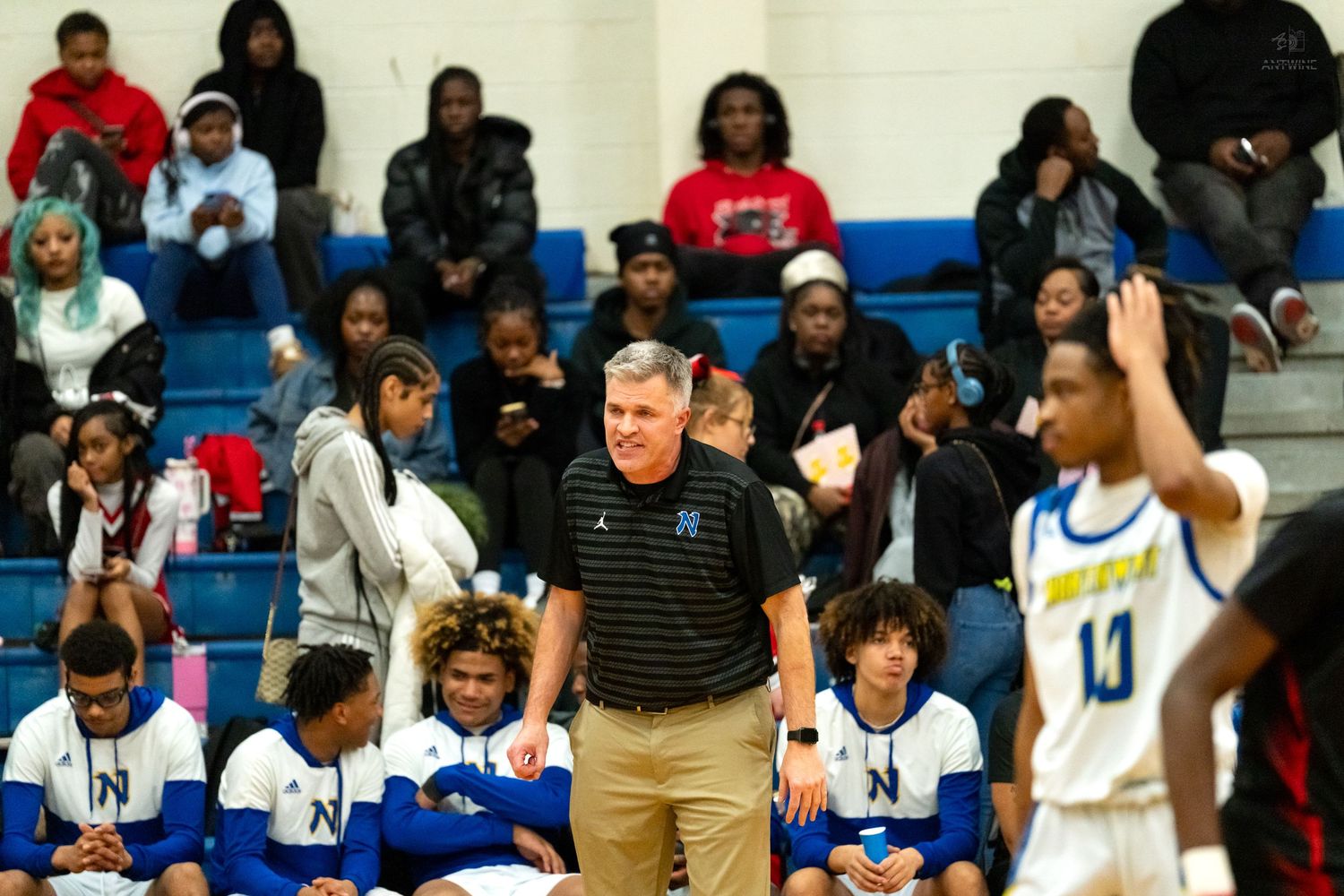 High school basketball coach smiles on the sideline during a game with players seated on the bench.