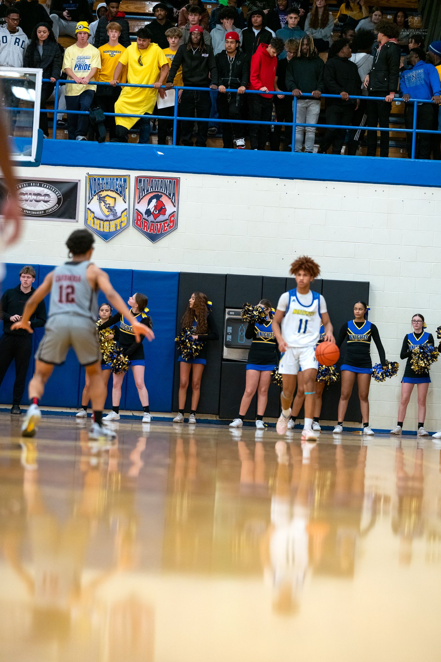 High school basketball player number 11 sets up play near half court with cheerleaders and crowd behind.