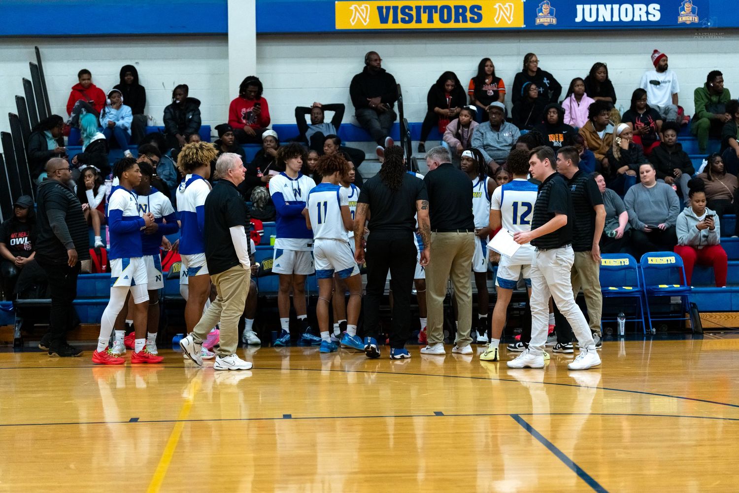 High school basketball team huddles with coaches during a timeout in a blue and gold gymnasium.