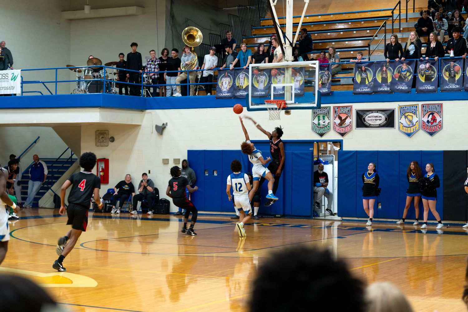 Basketball player in red uniform attempts layup against blue team defender in packed high school gymnasium.