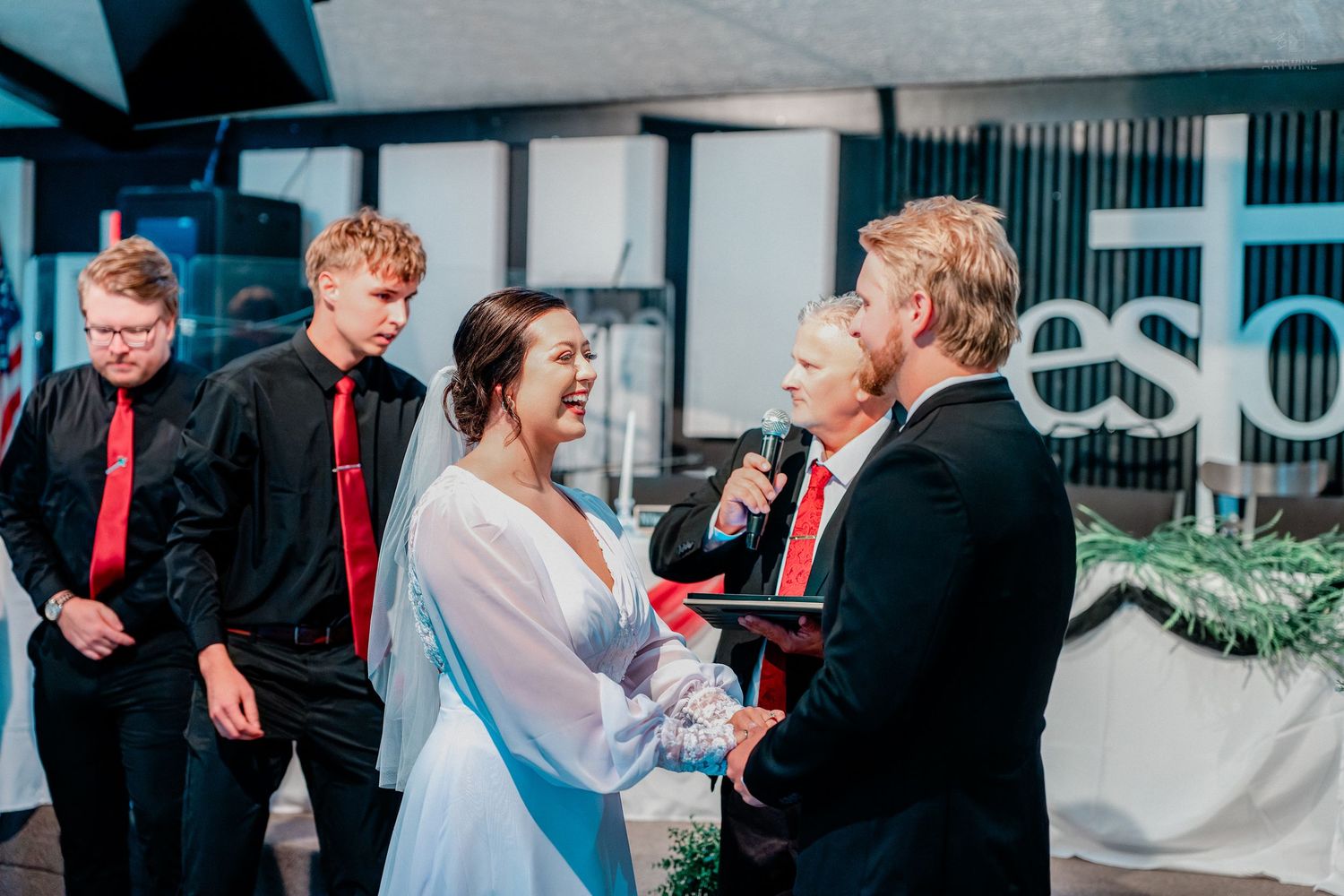 Bride and groom exchanging vows at wedding ceremony with groomsmen in red ties in modern venue.