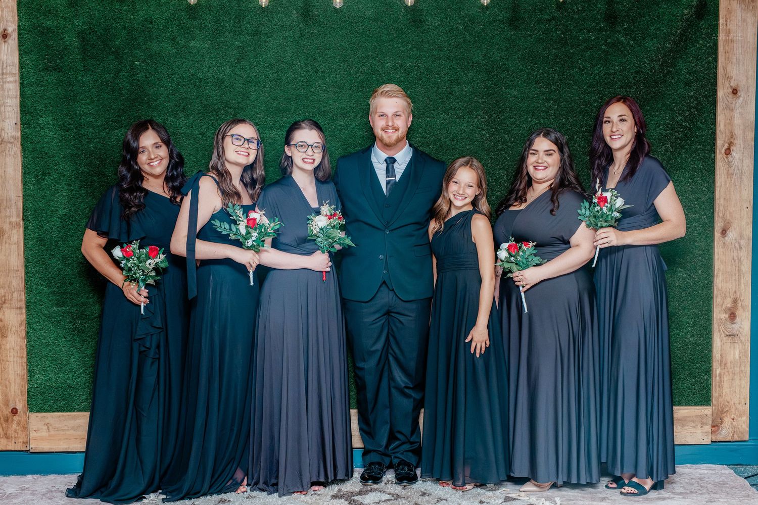 Groom in teal suit poses with six bridesmaids in navy dresses holding bouquets before a green hedge wall.