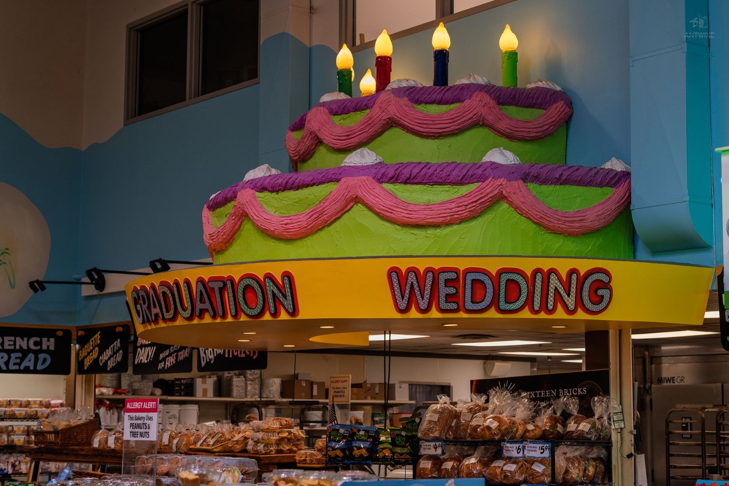 Giant cake decoration above bakery counter with Graduation and Wedding signs in a grocery store.