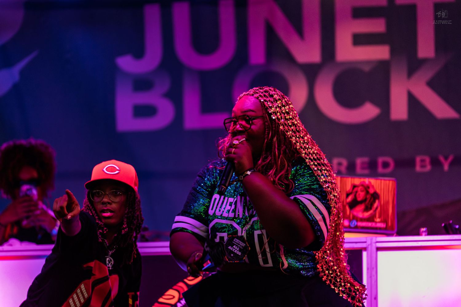 Two performers on stage at a Juneteenth Block Party event, with colorful lighting and a crowd in the background.