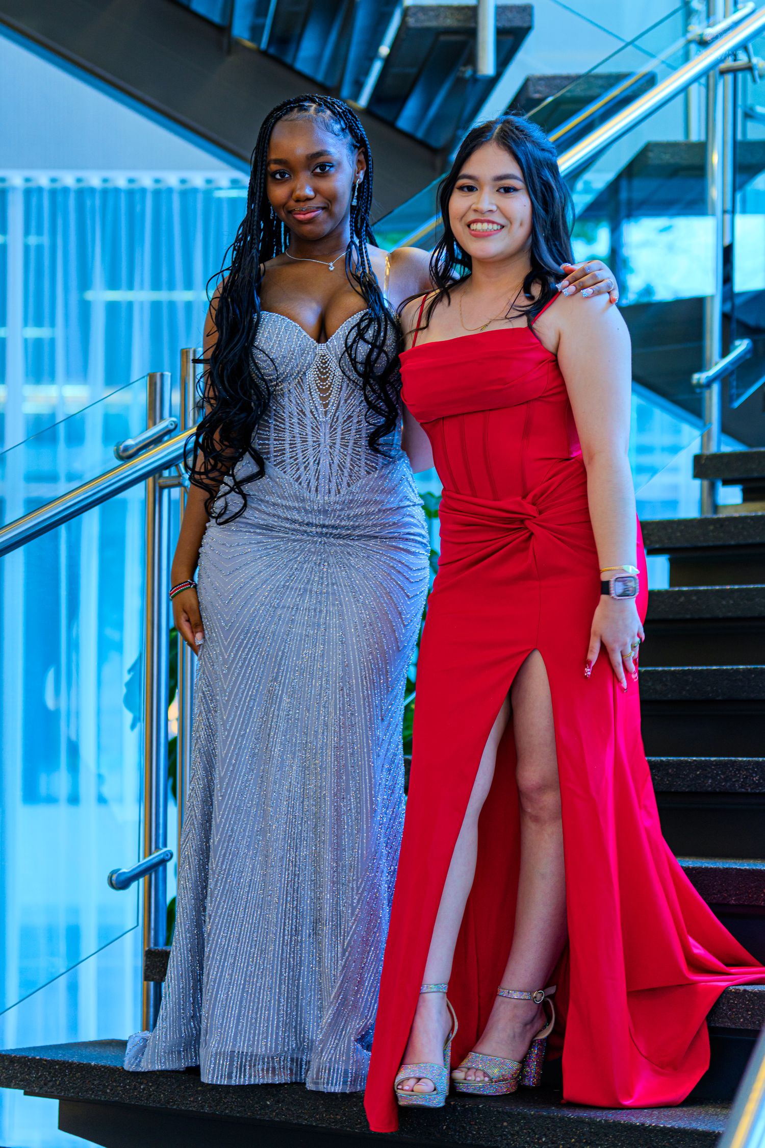 Two young women in elegant formal gowns, one silver and one red, posing on stairs at prom.