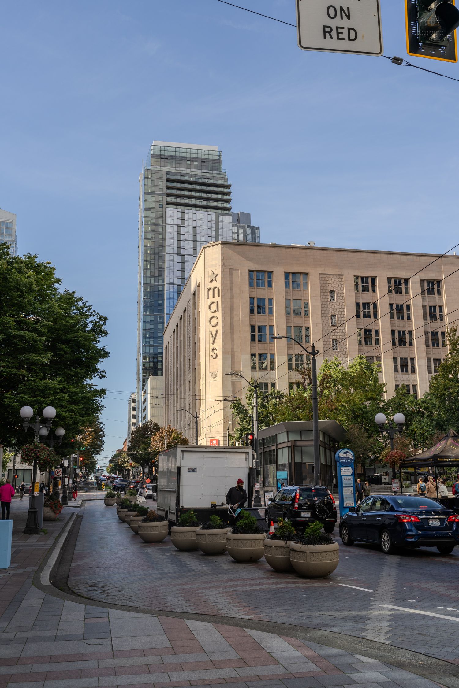 Macy's department store on a bustling Seattle street corner with modern skyscraper and tree-lined sidewalk.