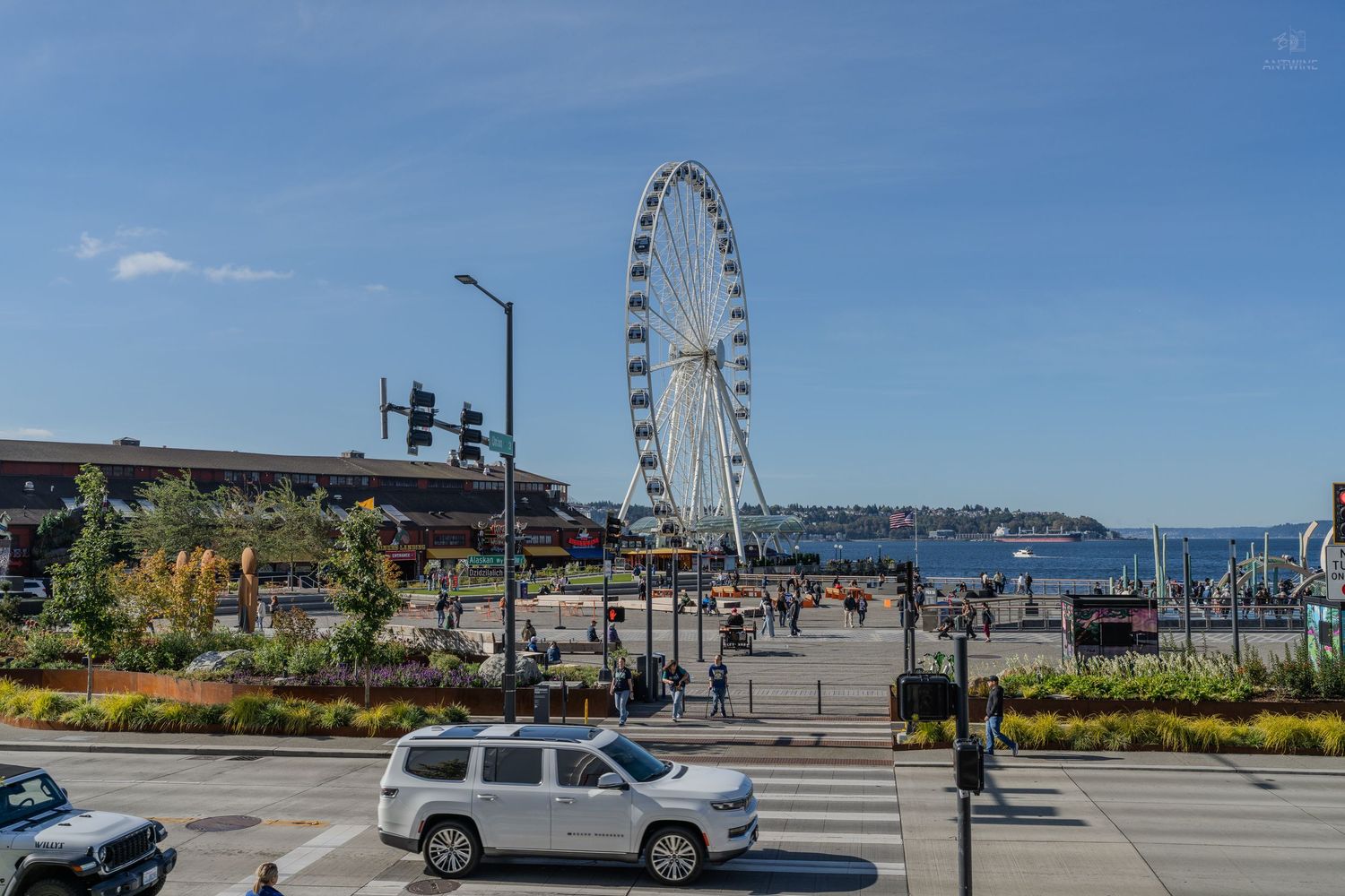 Seattle Great Wheel Ferris wheel at Pier 57 waterfront with white SUV crossing intersection on sunny day.