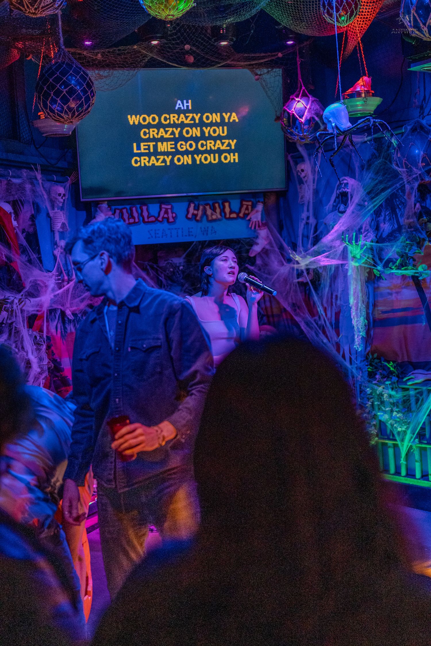 Woman singing Crazy on You karaoke at Halloween-decorated Hula Hula bar with blue neon lights.