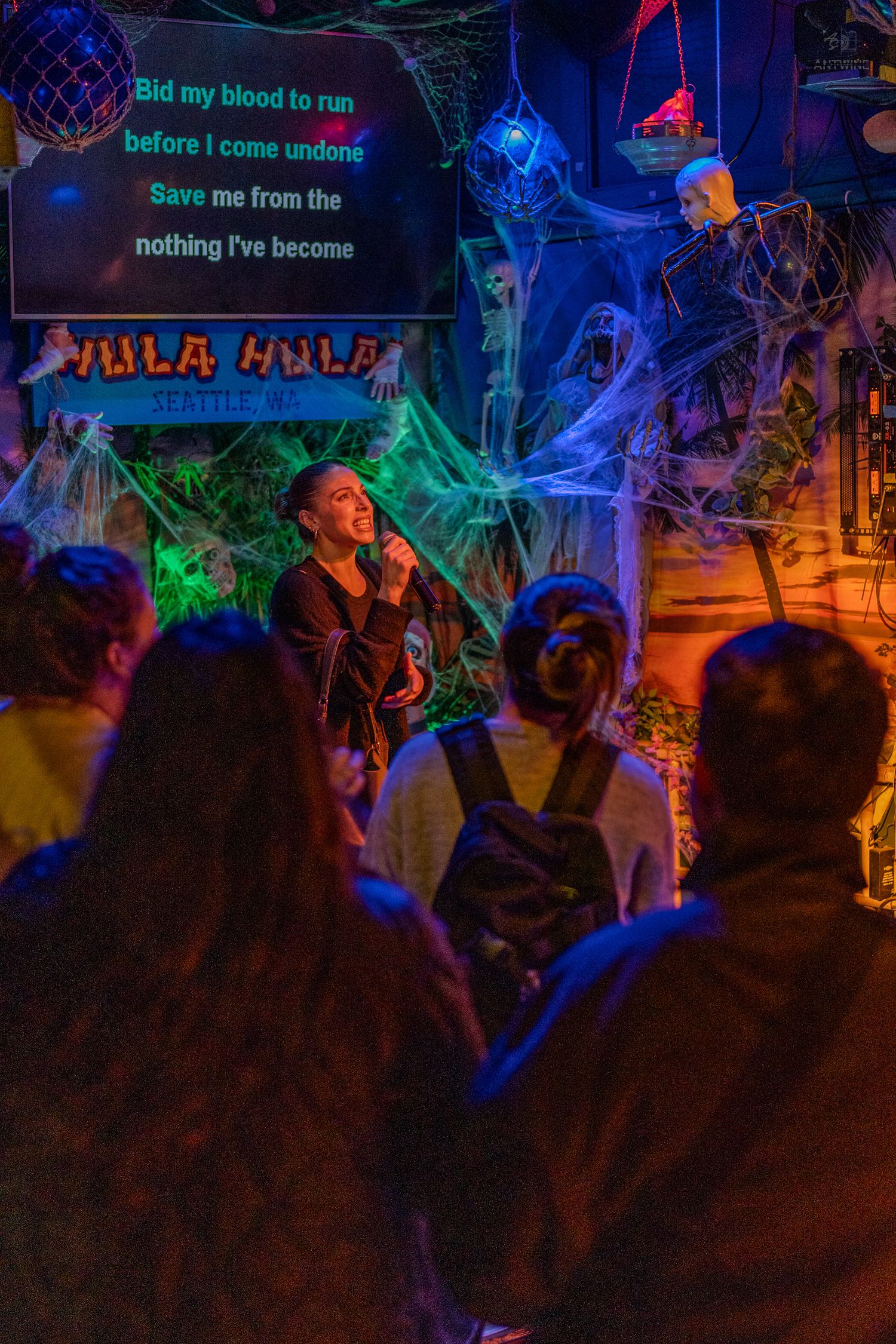 Woman singing karaoke on Halloween-themed stage before an audience at Hula Hula bar.