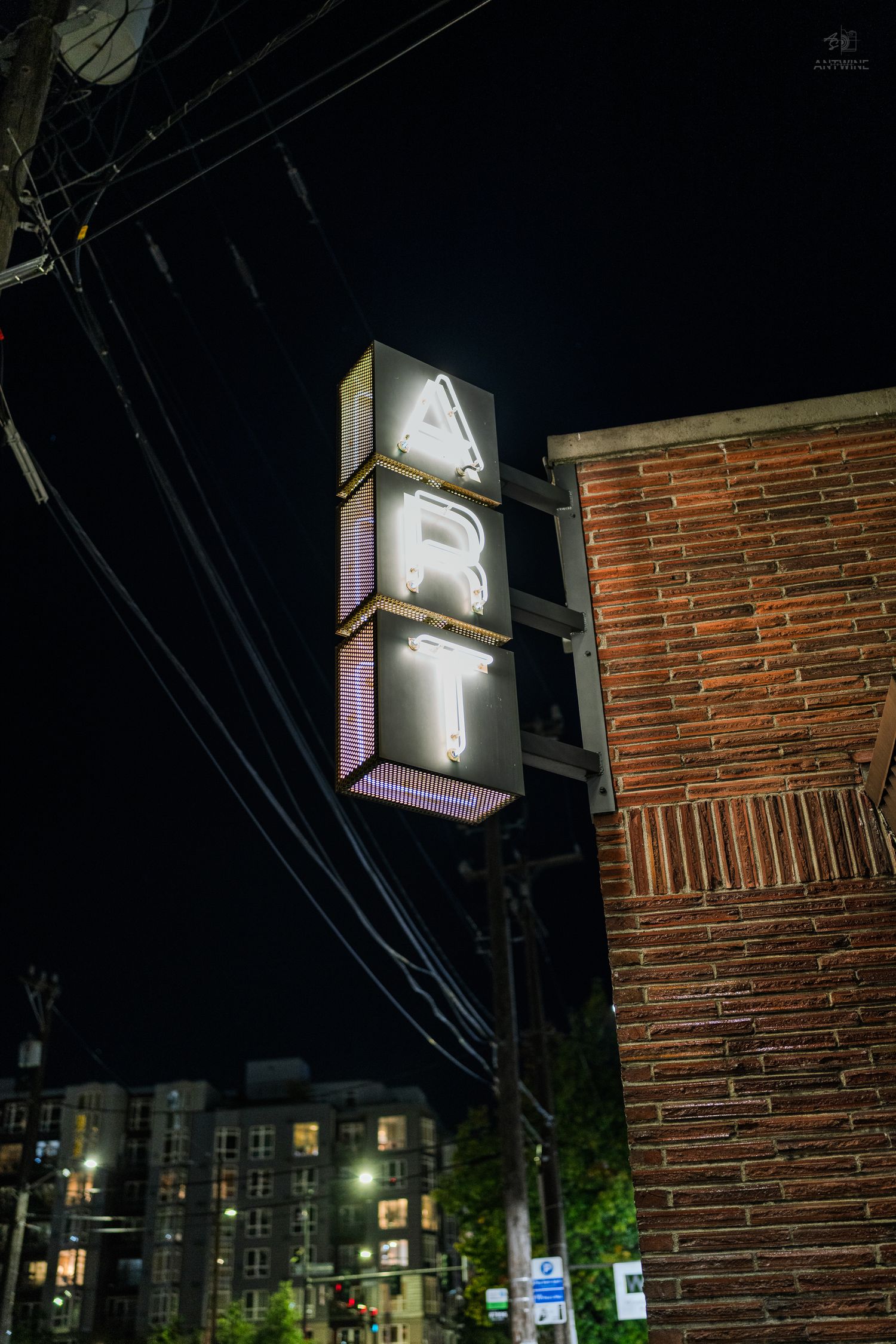 Illuminated vertical ART neon sign on brick building at night with city lights in background.