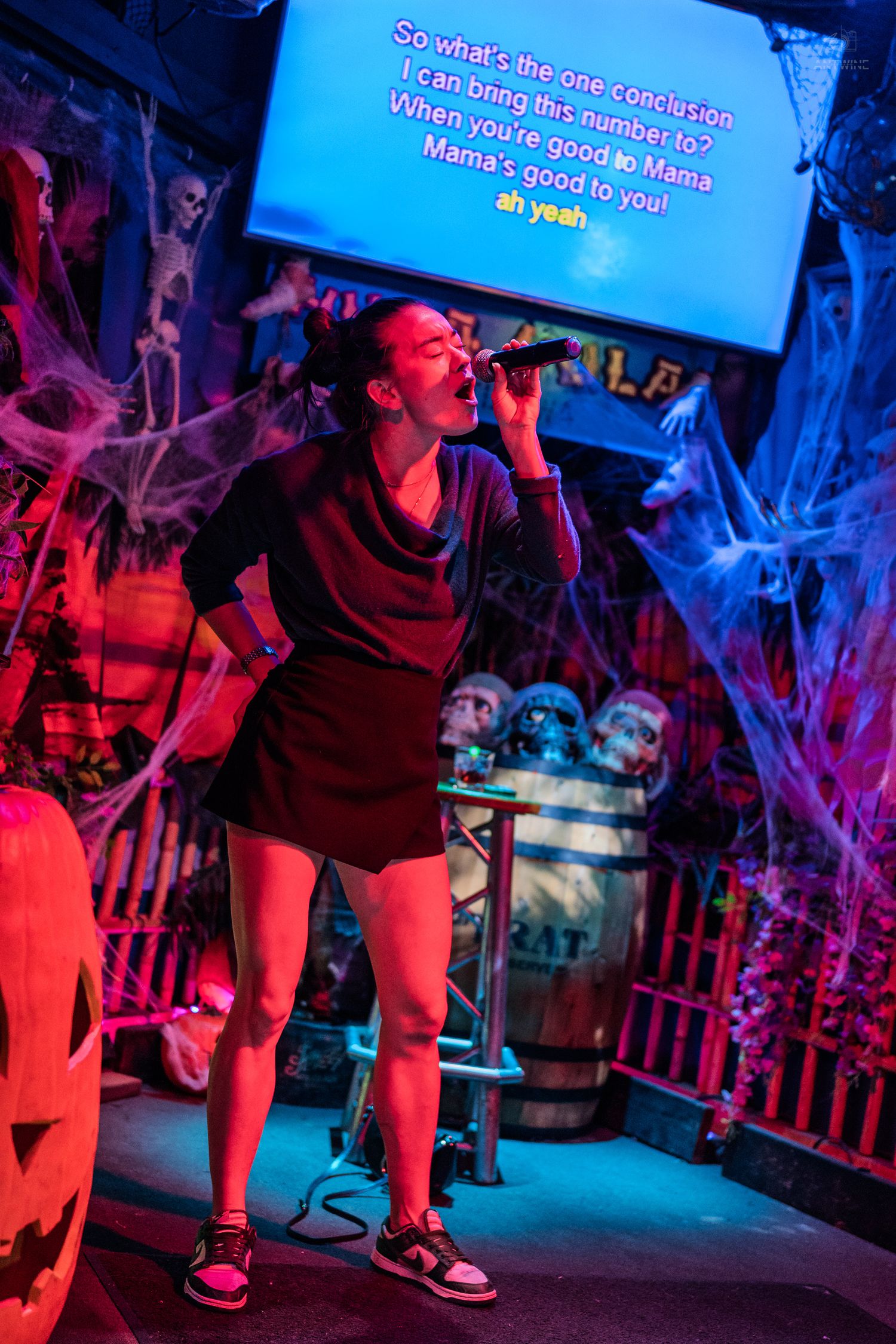 Woman singing karaoke on Halloween-decorated stage with spiderwebs, skulls, and pumpkins under colorful neon lights.