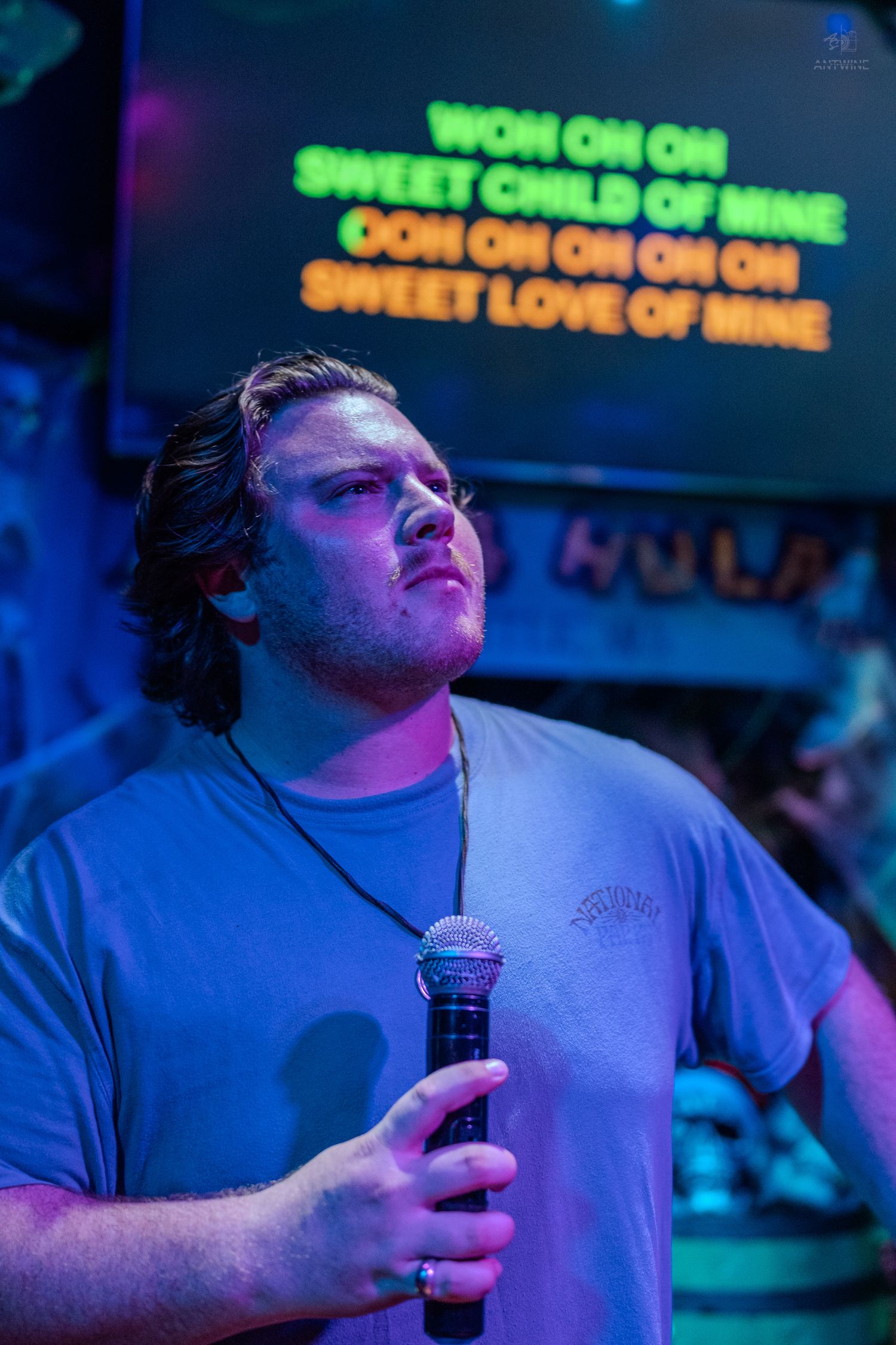 Man singing karaoke to Sweet Child of Mine by Guns N Roses, holding microphone under colorful stage lights.