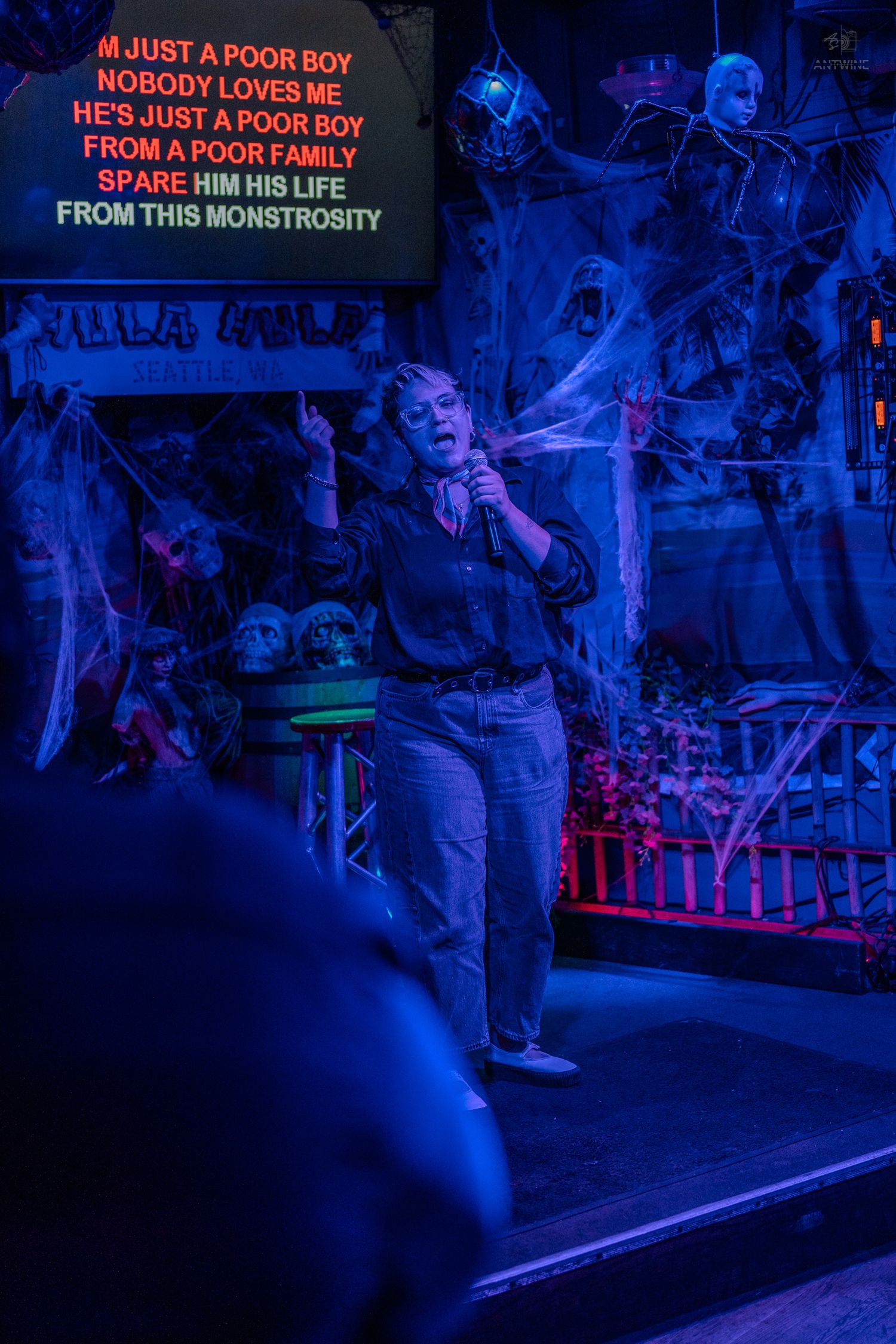 Woman performing karaoke on spooky Halloween stage with cobwebs, skulls, and blue lighting, singing Bohemian Rhapsody.