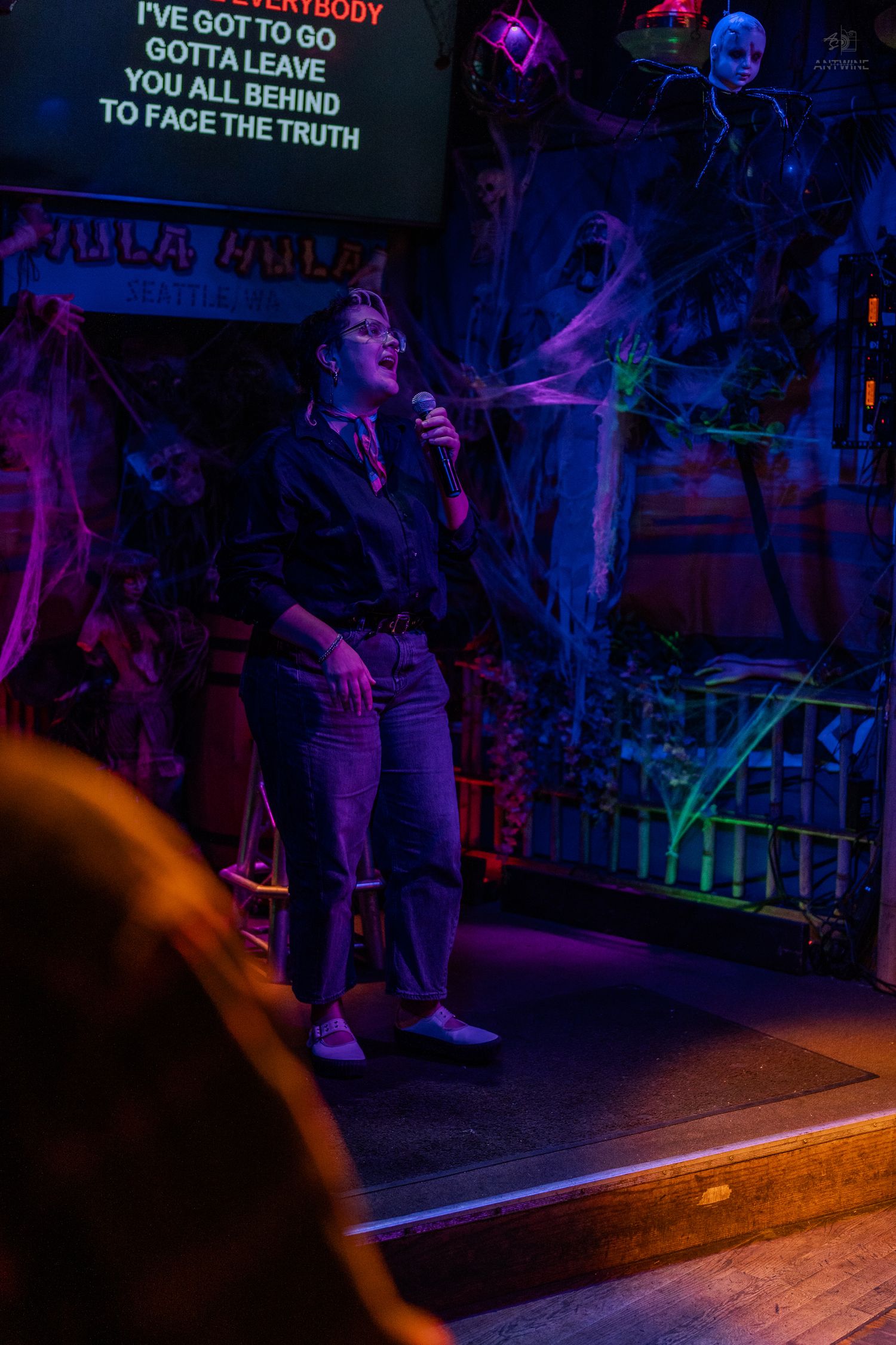 Person singing karaoke on a Halloween-decorated stage with colorful neon lights and spooky decorations.