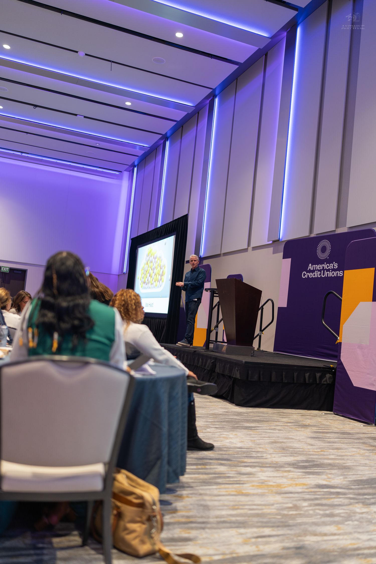 Attendees listen to a presenter at an America's Credit Unions conference in a modern ballroom with purple LED lighting.
