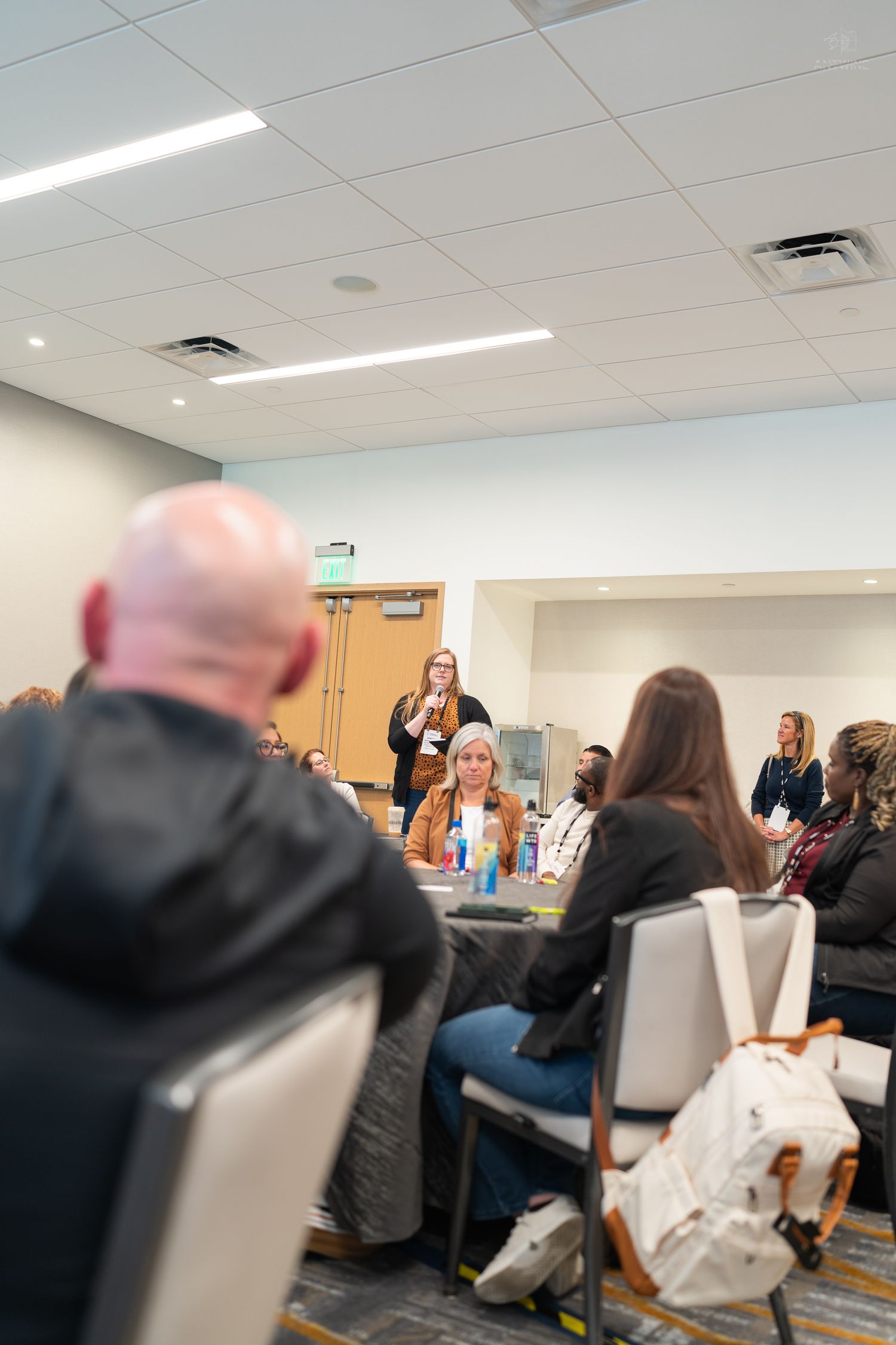 Woman speaking at a podium during an indoor conference session with attendees seated at round tables.