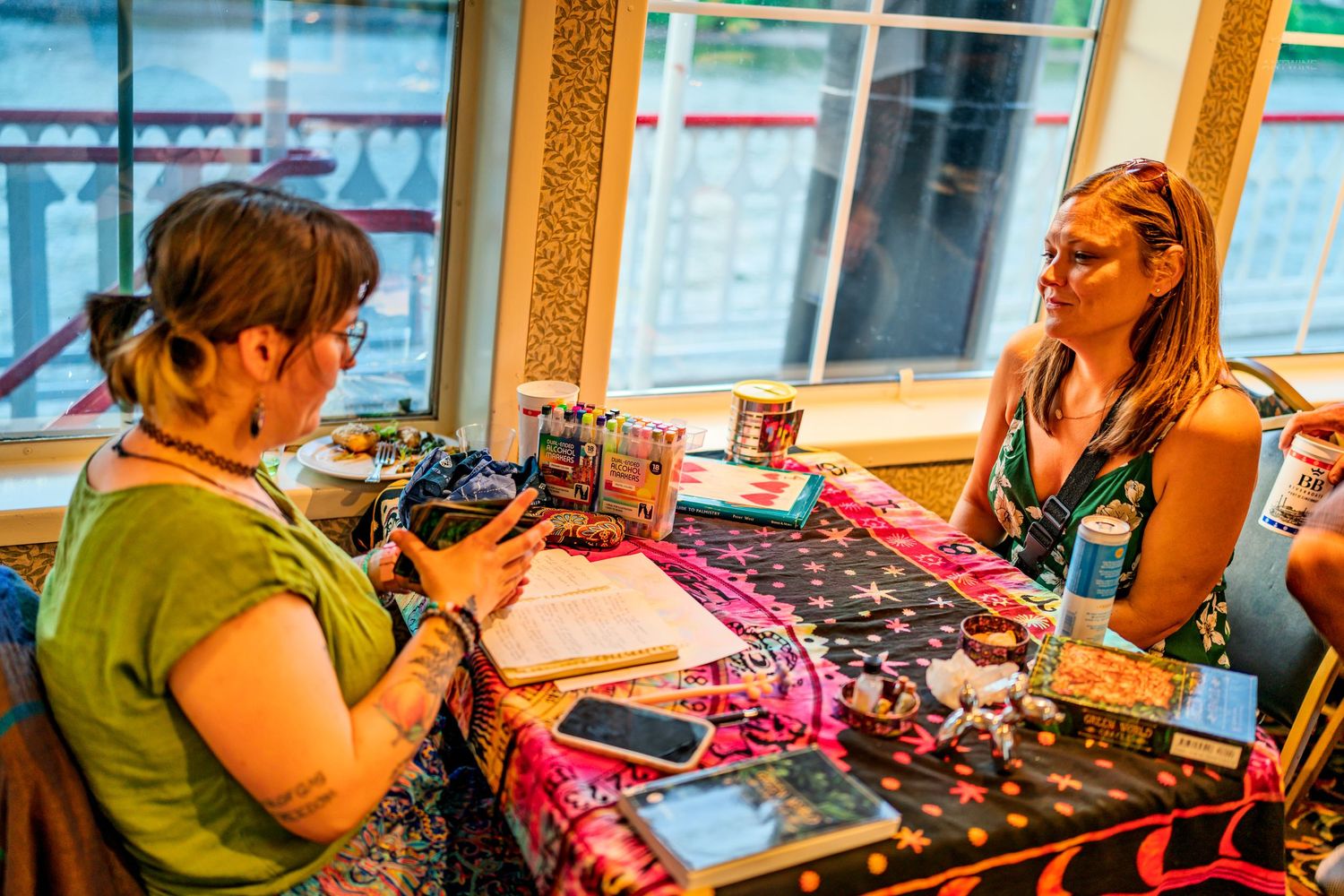 Two women sitting across table covered with craft supplies near bright window.