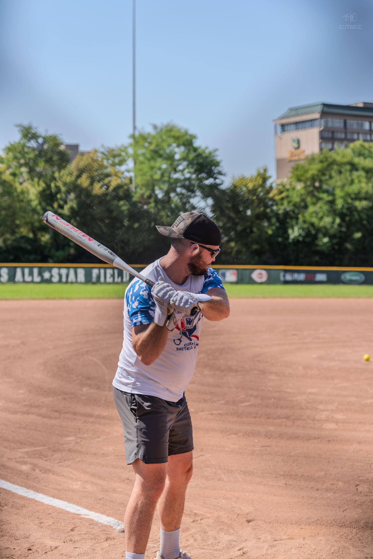 Man in cap and sunglasses holding a baseball bat on a sunny softball field.