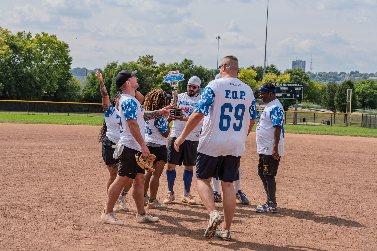 FOP softball team players celebrating on the field during a sunny outdoor game on a dirt infield.