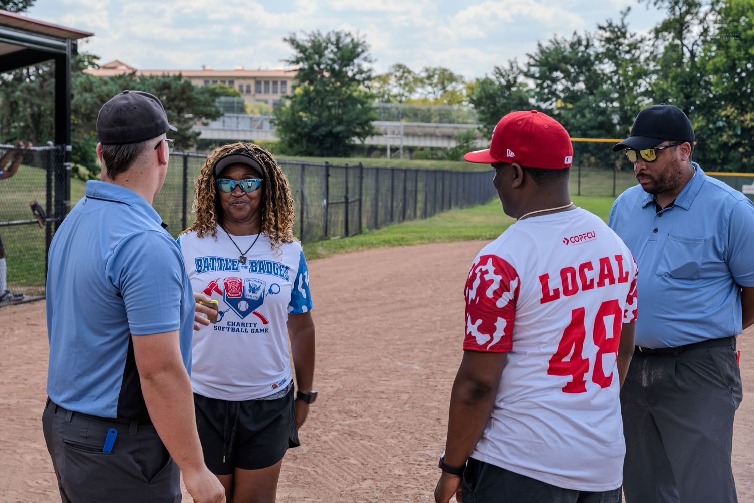 Two umpires meeting with softball team captains in FOP and Local 48 jerseys on a dirt field.