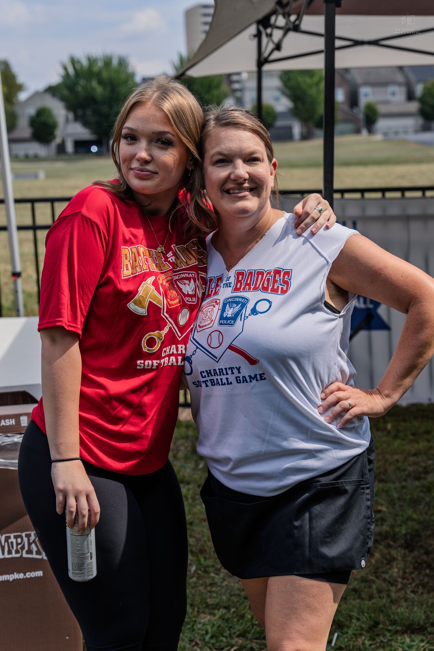 Two women posing together at a Battle of the Badges charity softball game event outdoors.