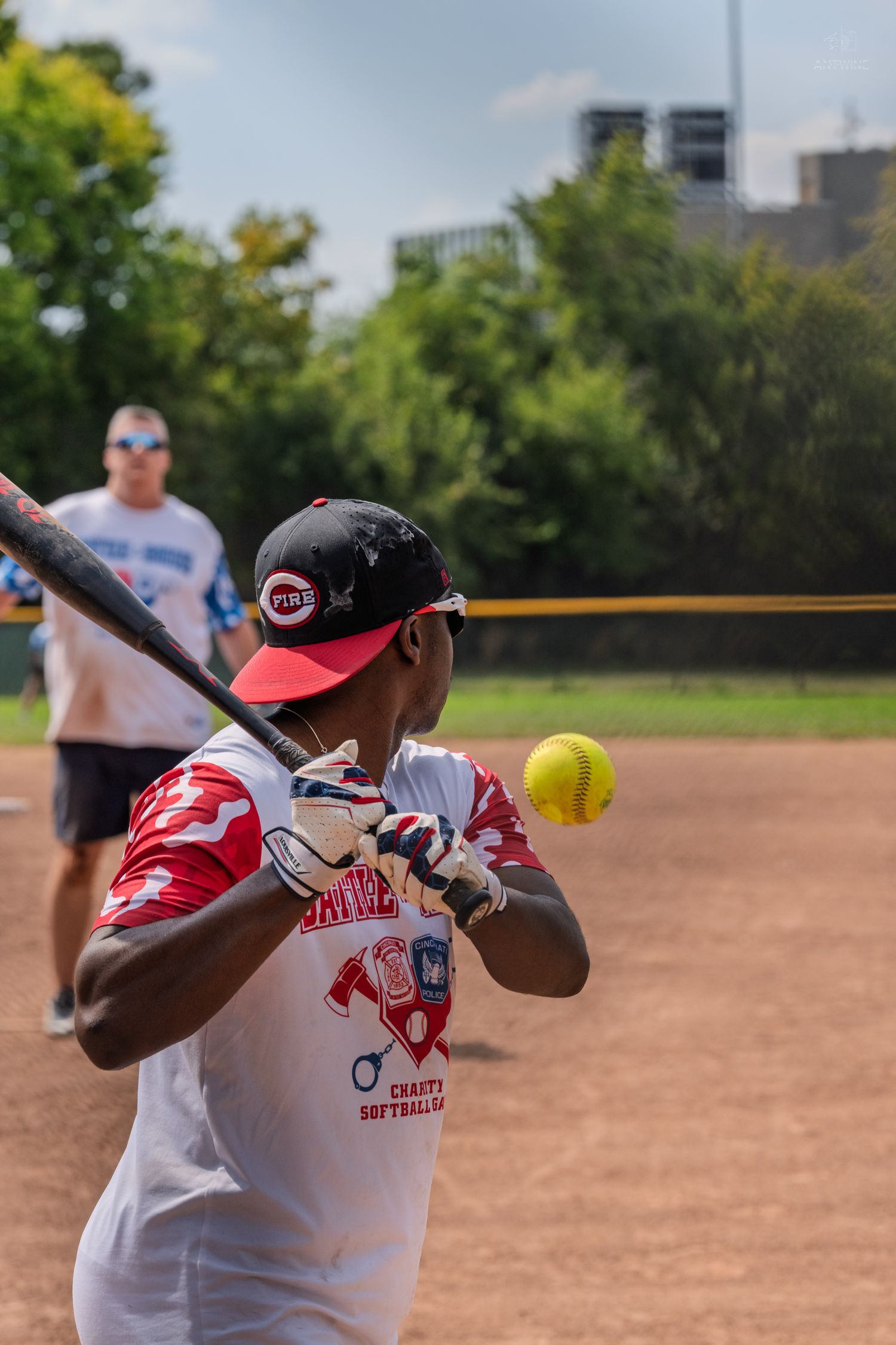 Close-up of batter in Cincinnati Reds cap swinging at yellow softball during charity softball game.