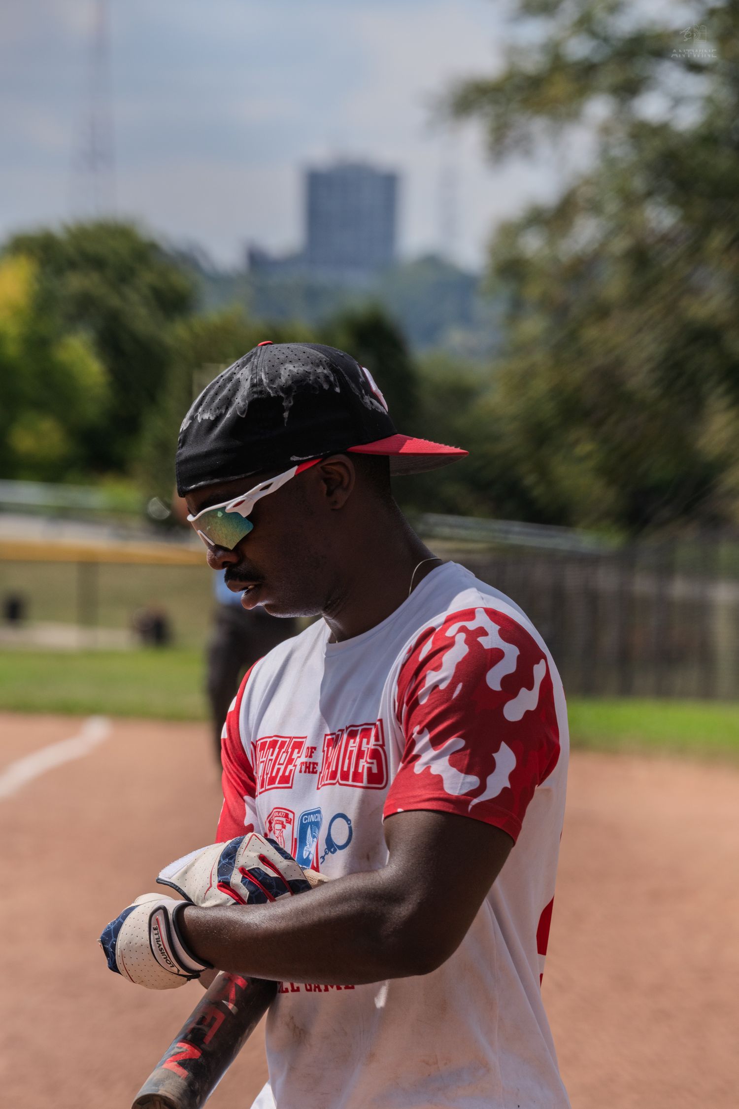 Baseball player in red camo jersey and sunglasses holding a bat at a charity softball game outdoors.