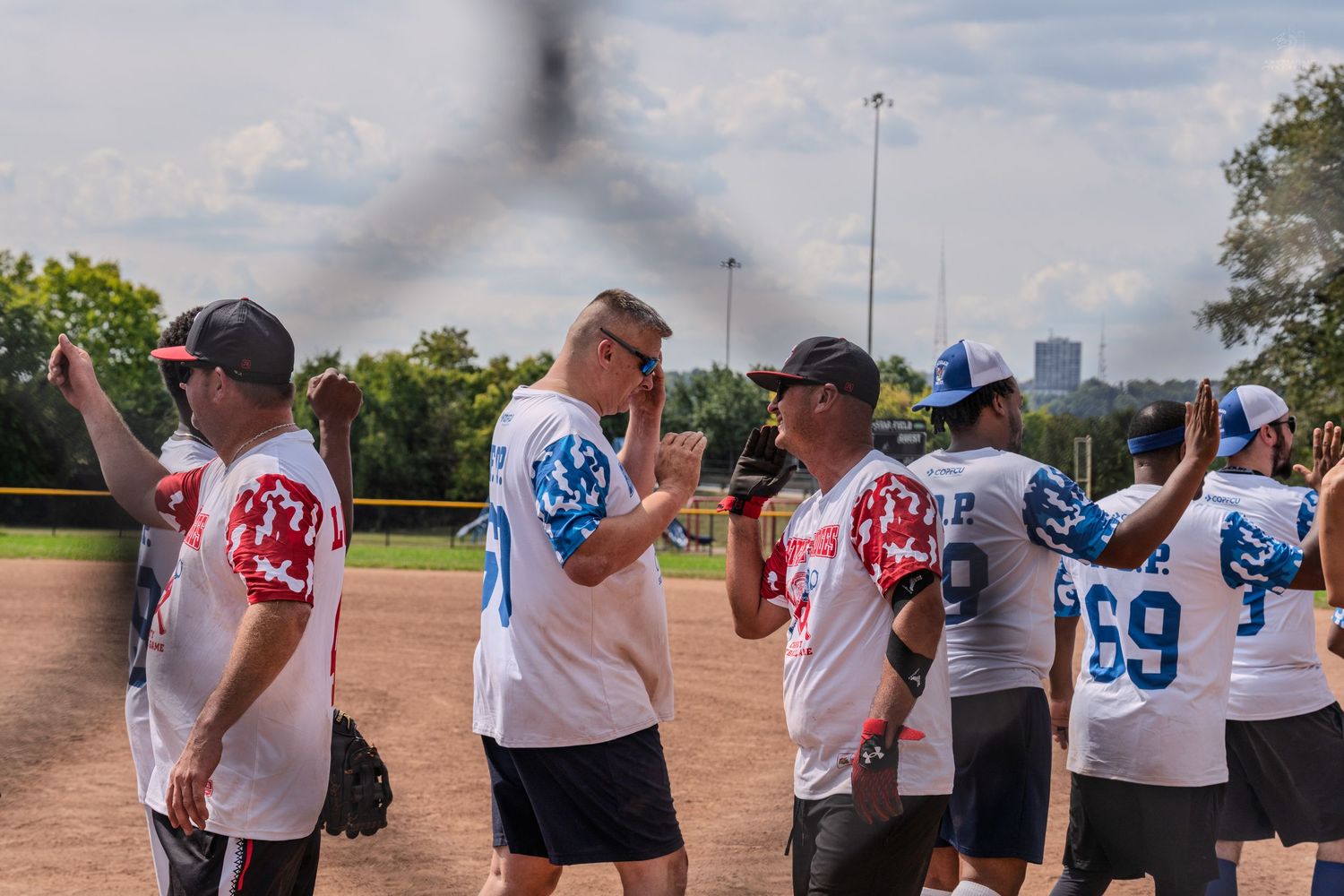 teammates in white patriotic jerseys celebrate on a softball field during an outdoor game on a sunny day.