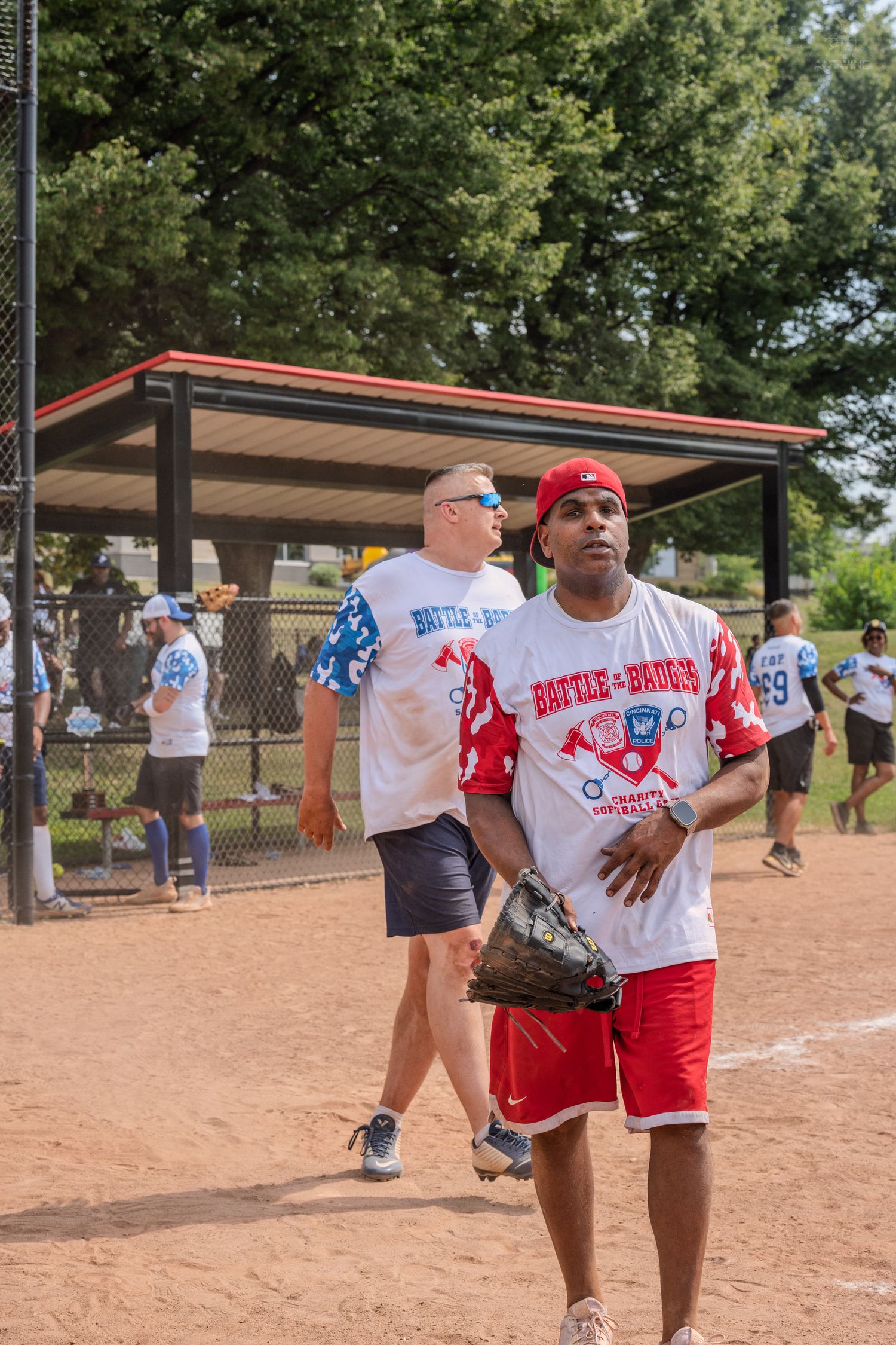 A player in a Battle of the Badges softball jersey holds a glove on a sunny baseball field.