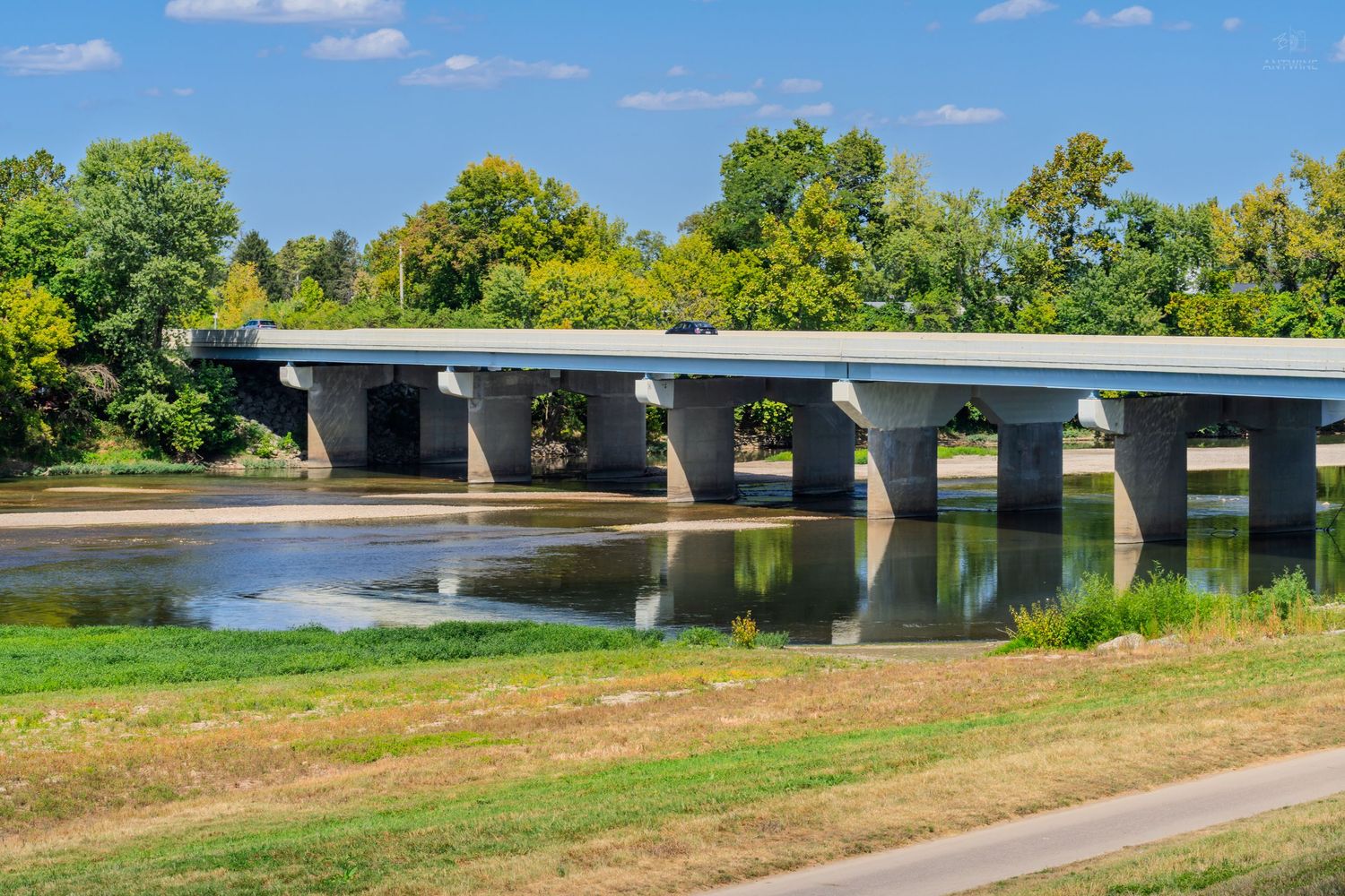 Concrete bridge with multiple pillars spanning a calm river, surrounded by lush green trees under a clear blue sky.