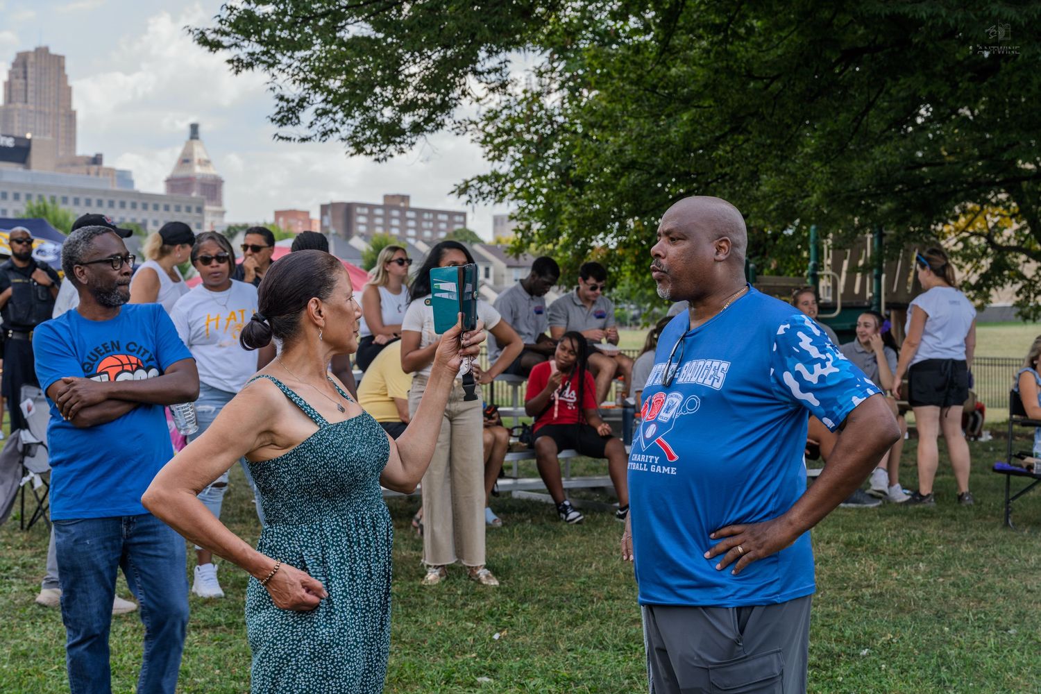 People gathered in a park for an outdoor community event with city buildings visible in the background.