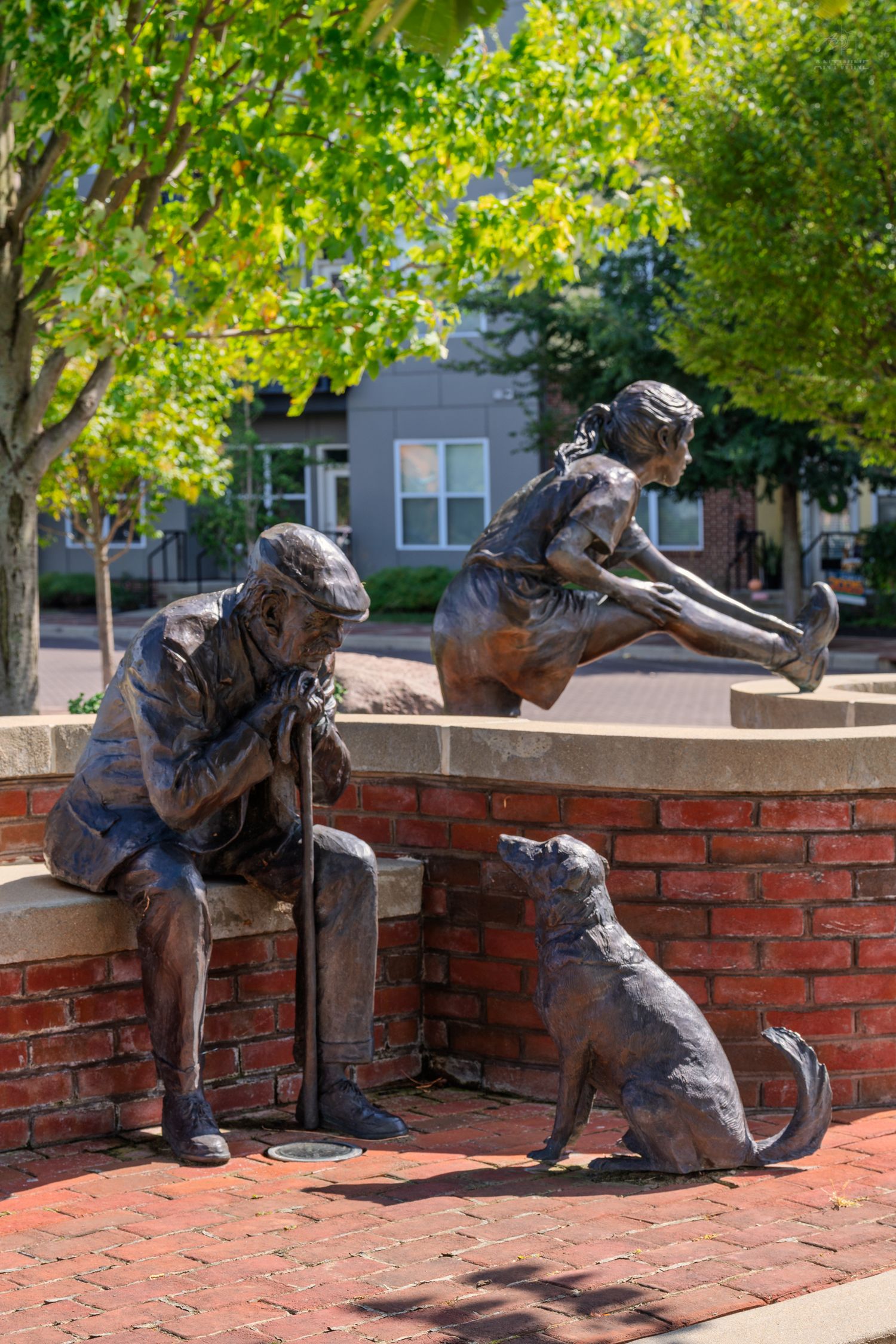 Bronze sculpture of elderly man, girl, and dog at brick fountain in park setting with green trees.