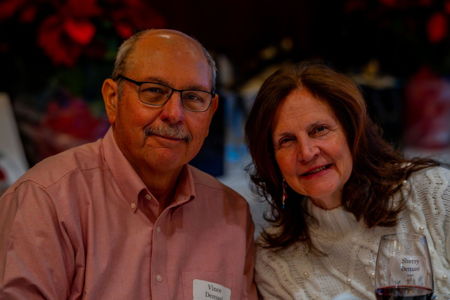 Smiling older couple posing together at a festive indoor event, man wearing glasses and pink shirt with name tag.
