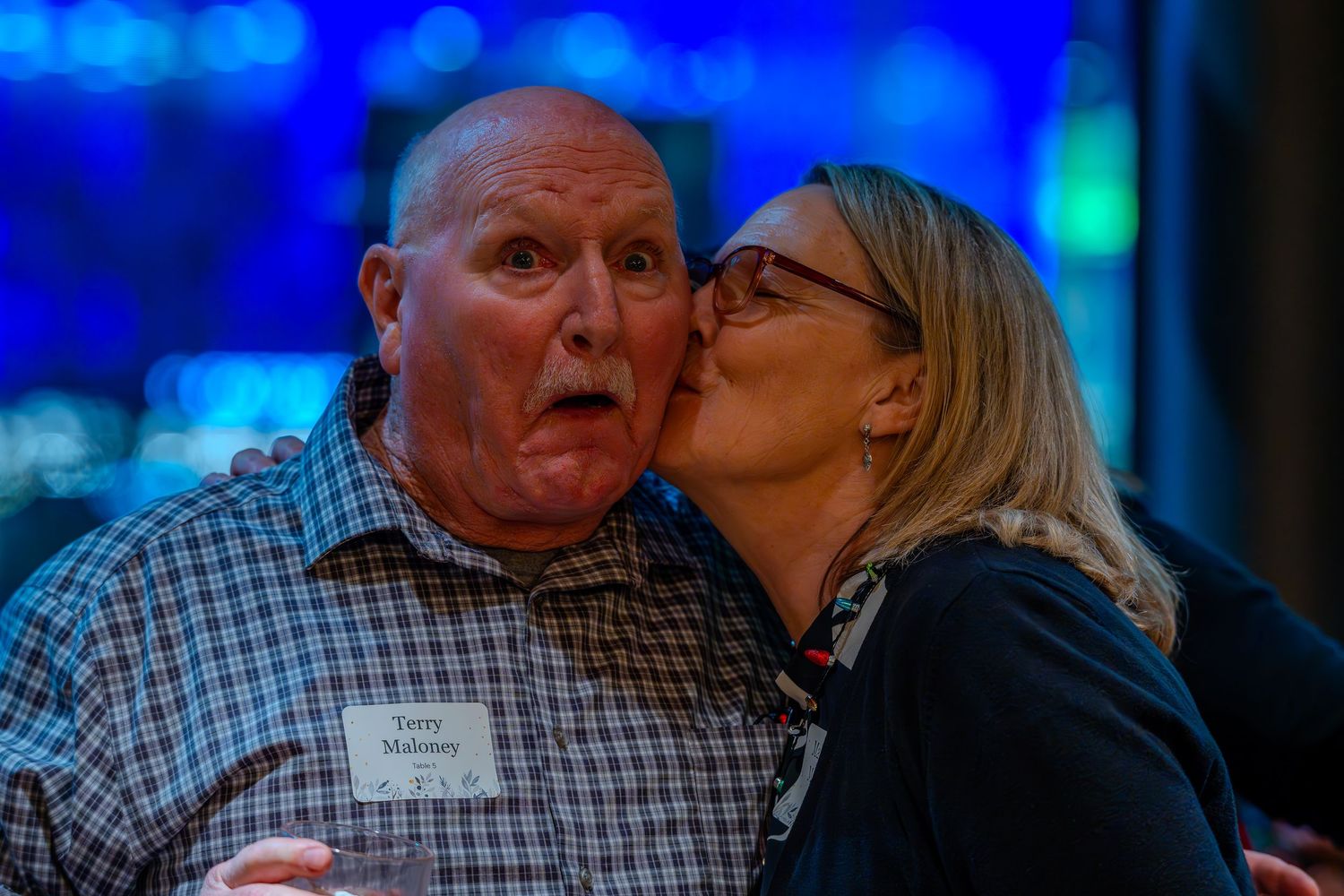 A woman kisses an older man on the cheek at a social event, both wearing name tags.