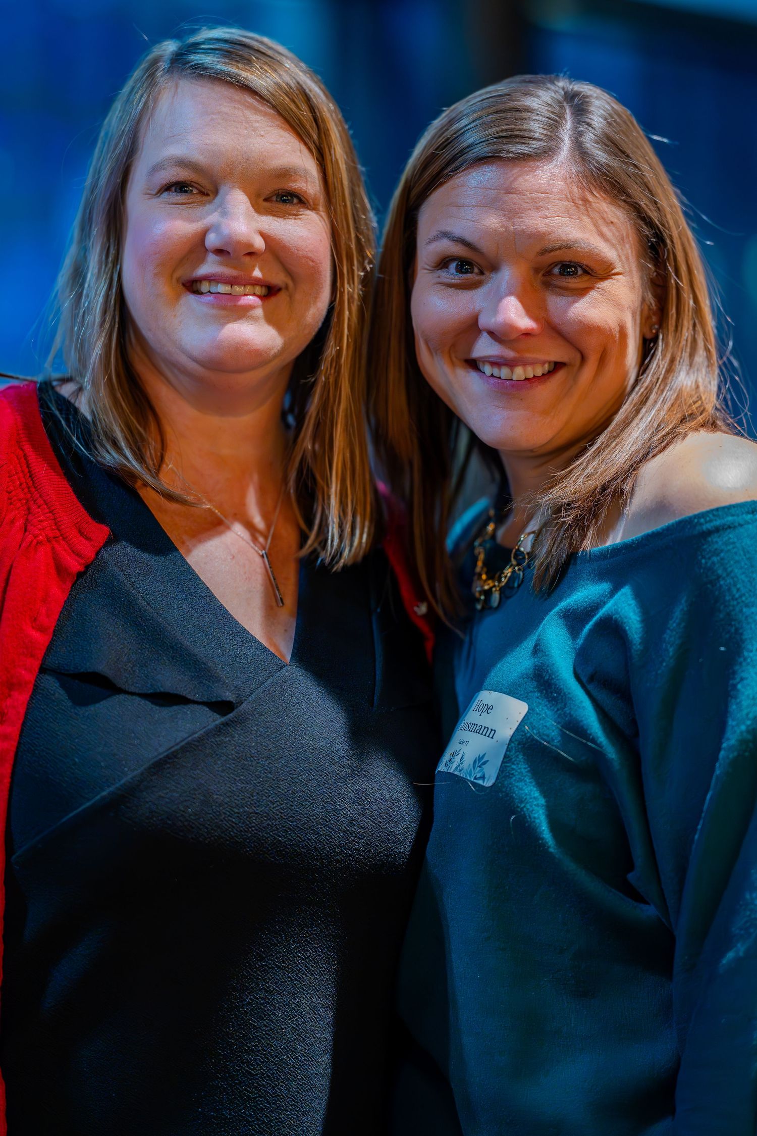 Two smiling women pose together at a formal event, one in black and one in teal, against a blue background.