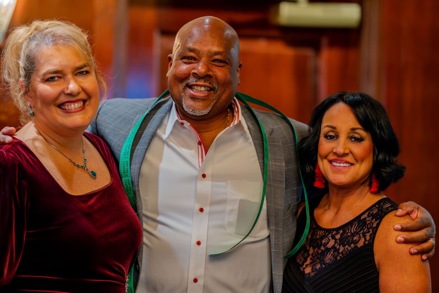 Three smiling adults posing together at a formal event, wearing elegant attire including velvet and lace outfits.