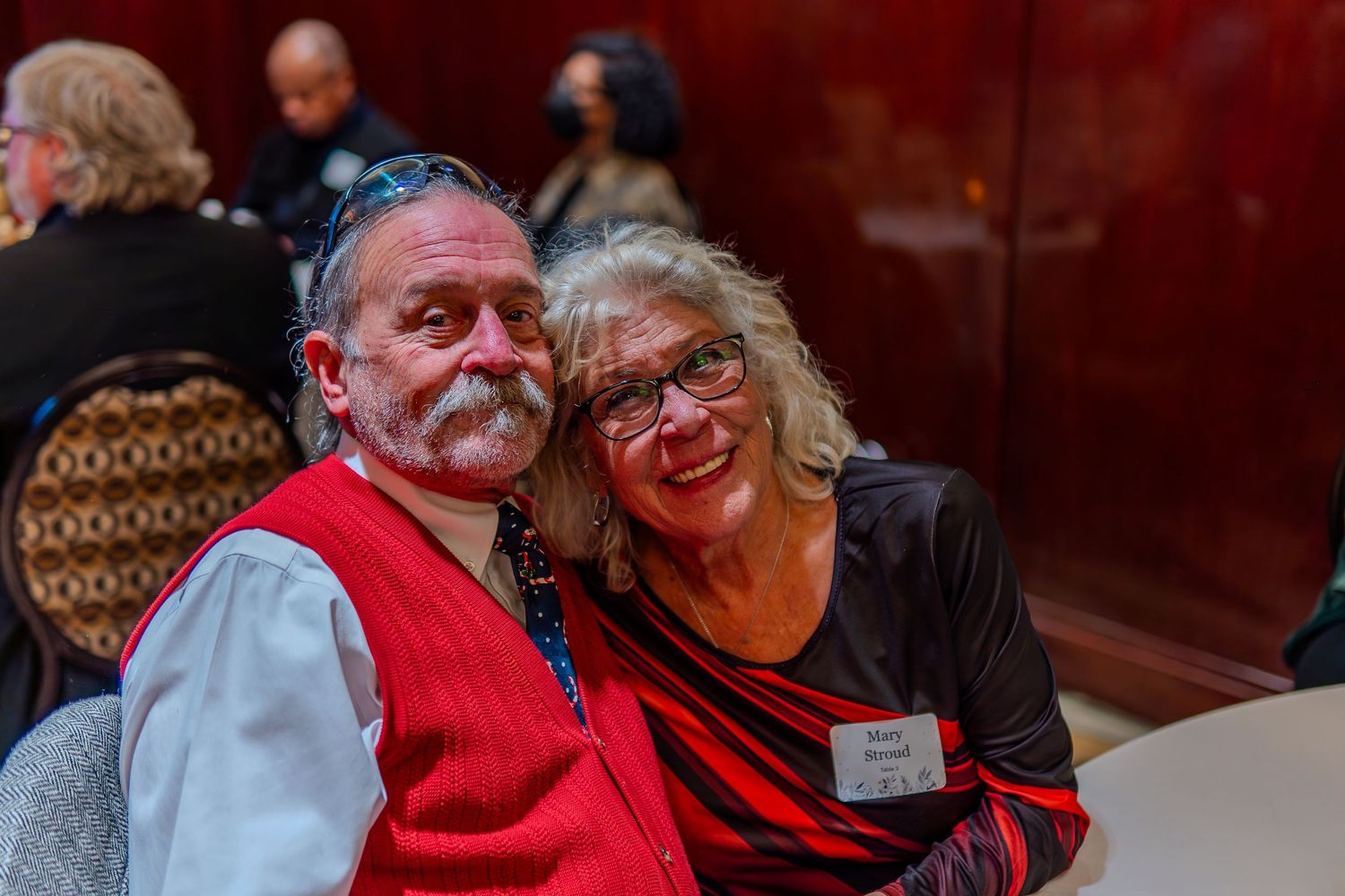 Smiling couple posing together at an indoor event, man in red vest and woman in black and red top wearing a name tag.