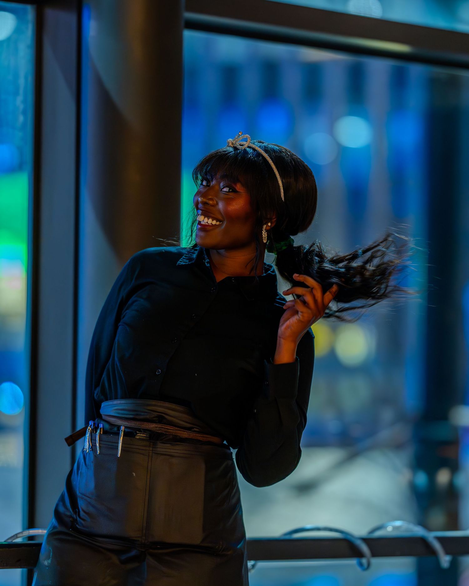 Smiling Black woman in black outfit and tiara headband holds feathered accessory indoors with blue bokeh background.