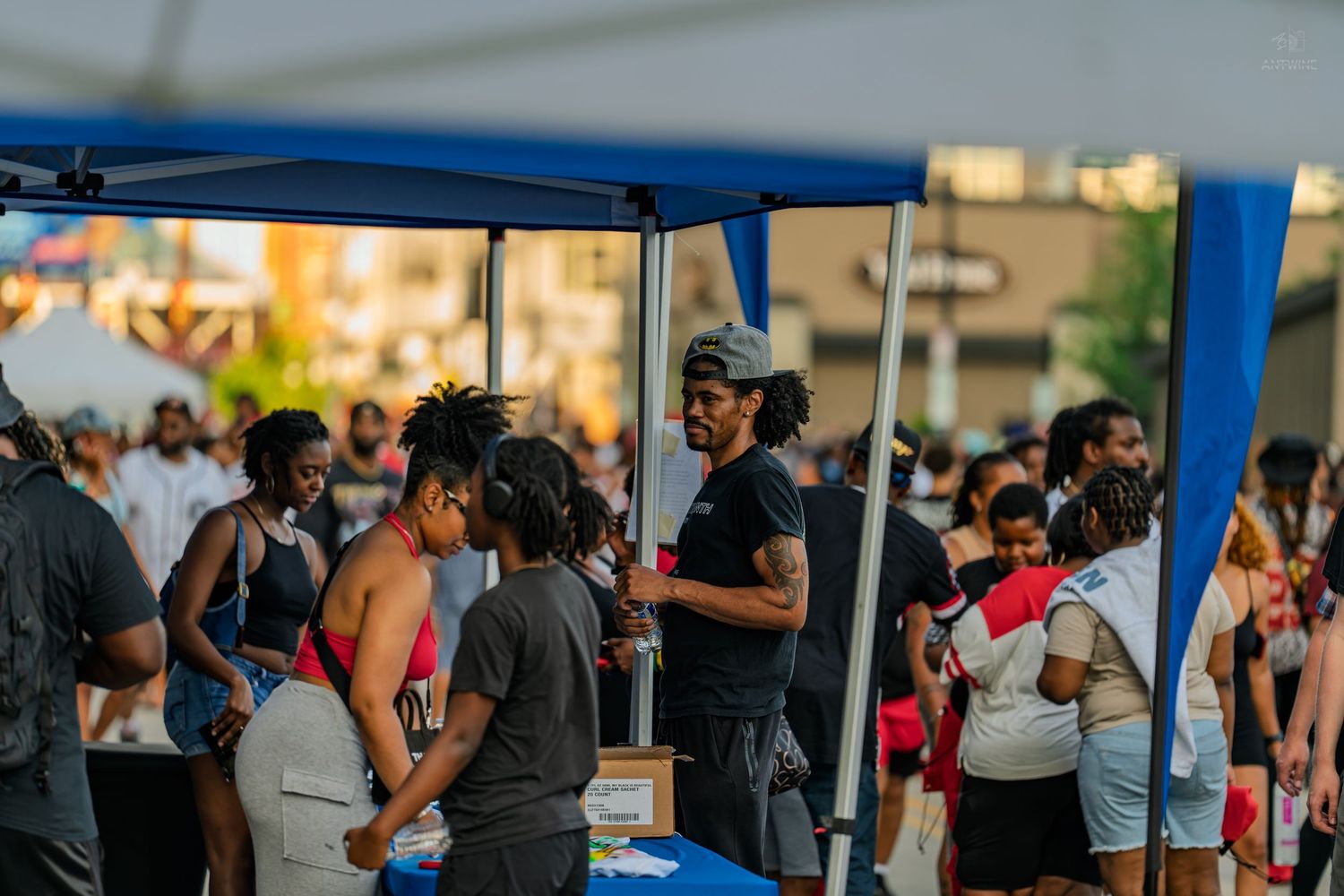Diverse crowd gathered at an outdoor community event under blue tent canopies at sunset.