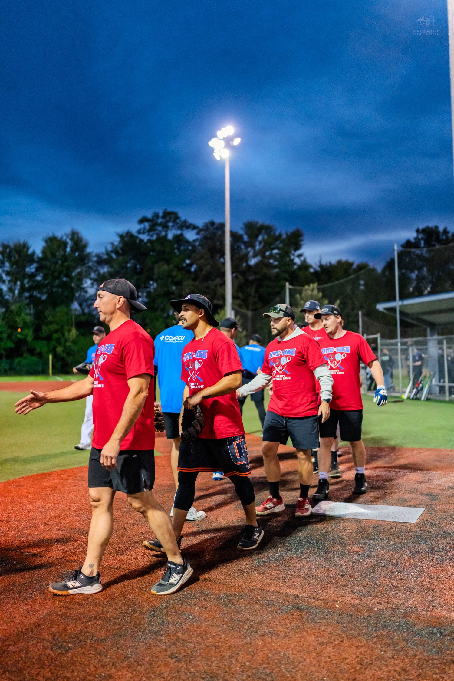 Softball team in matching red shirts walks together on field under bright stadium lights at night.