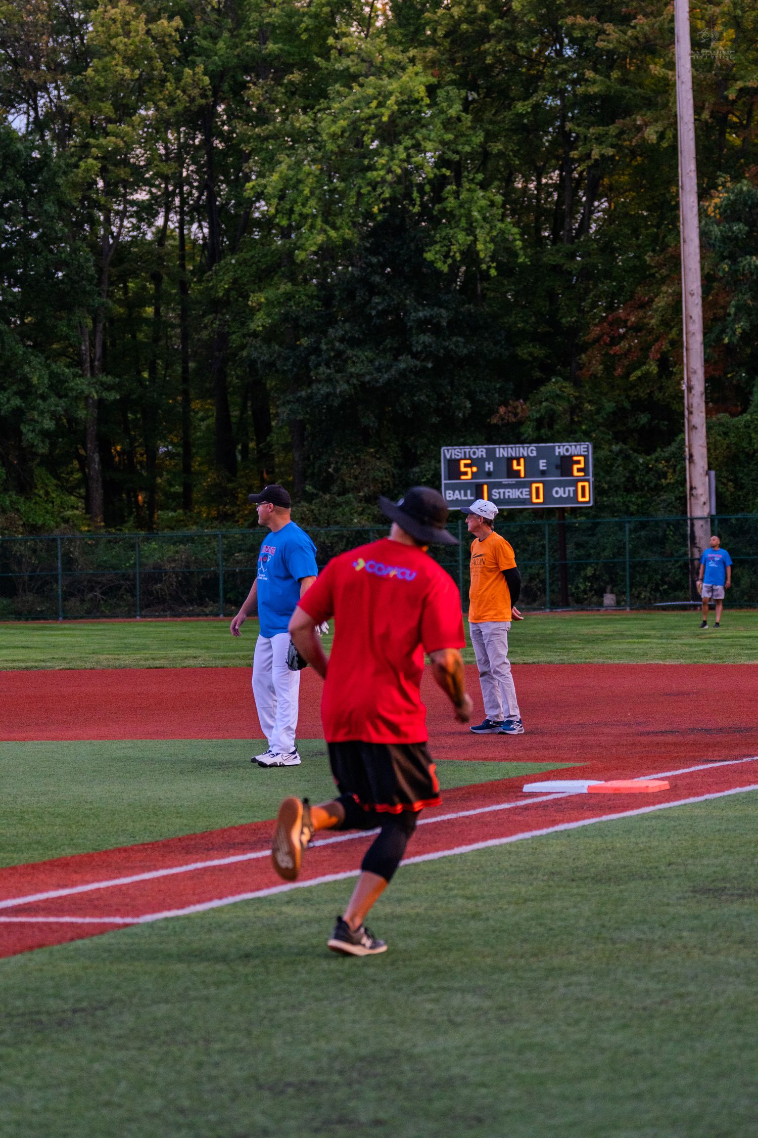 A baseball player in a red shirt runs the bases during an evening recreational league game with a scoreboard visible in the background.