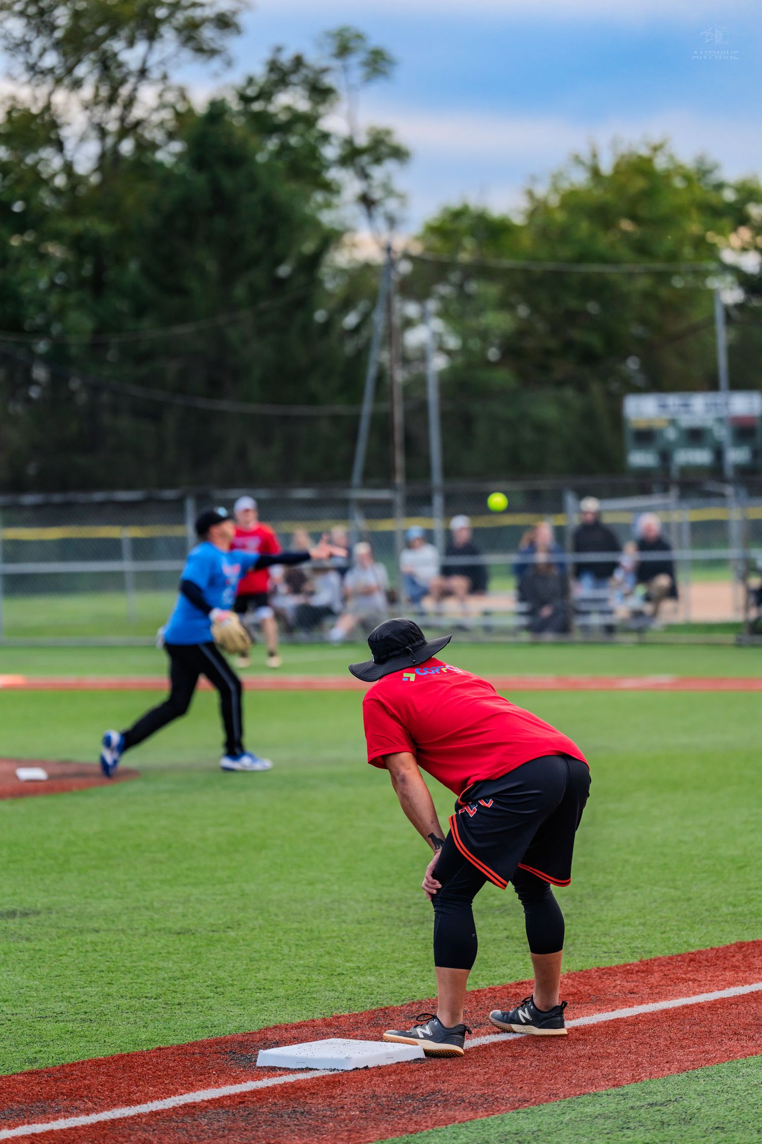 Softball player in red shirt ready at first base as pitcher releases yellow ball toward home plate.