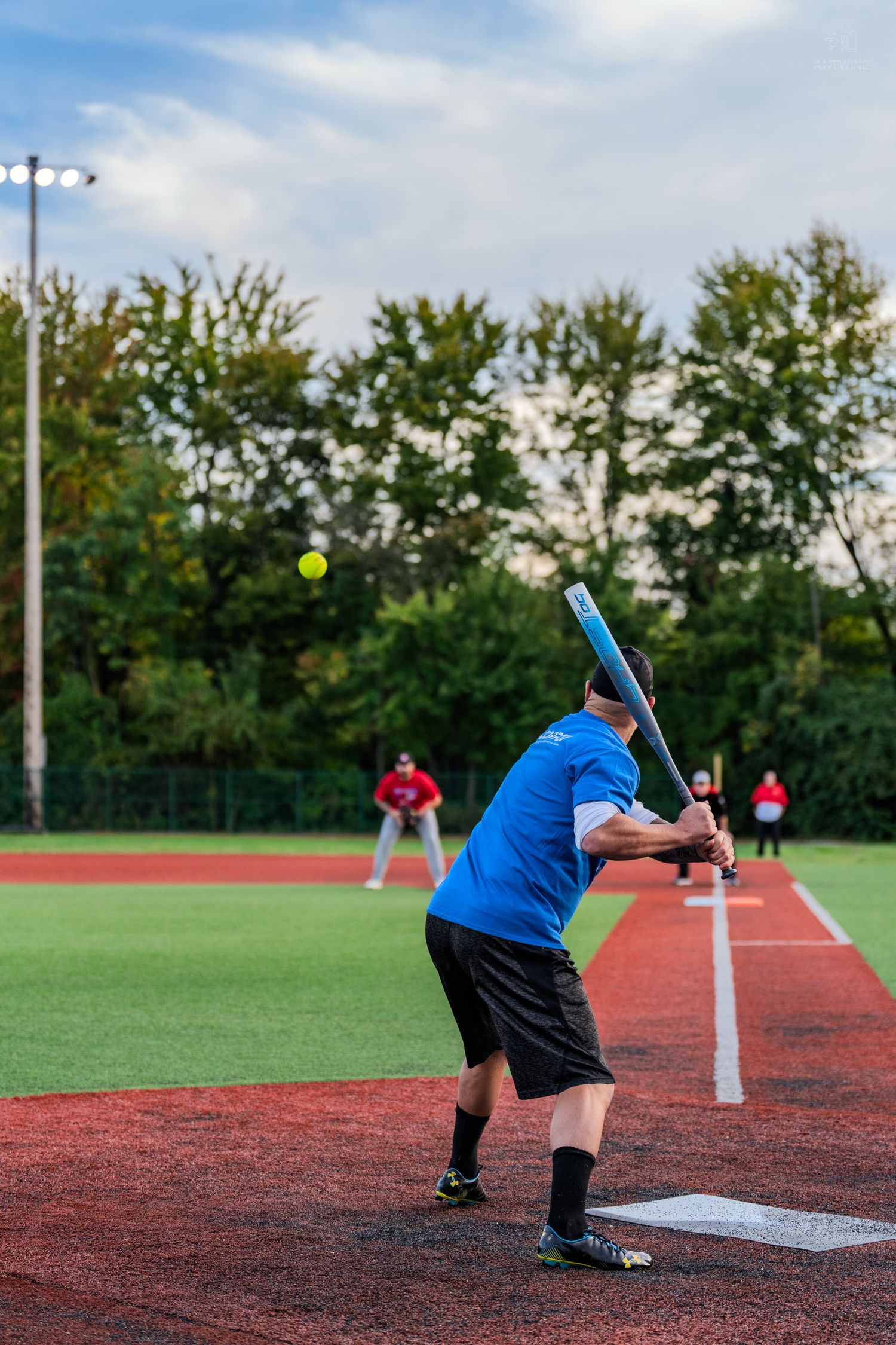 Softball batter in blue shirt swinging at yellow ball during outdoor game on red turf field.