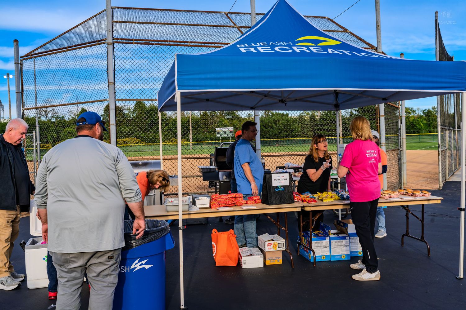 Recreation department tent with staff serving refreshments at outdoor baseball field event.
