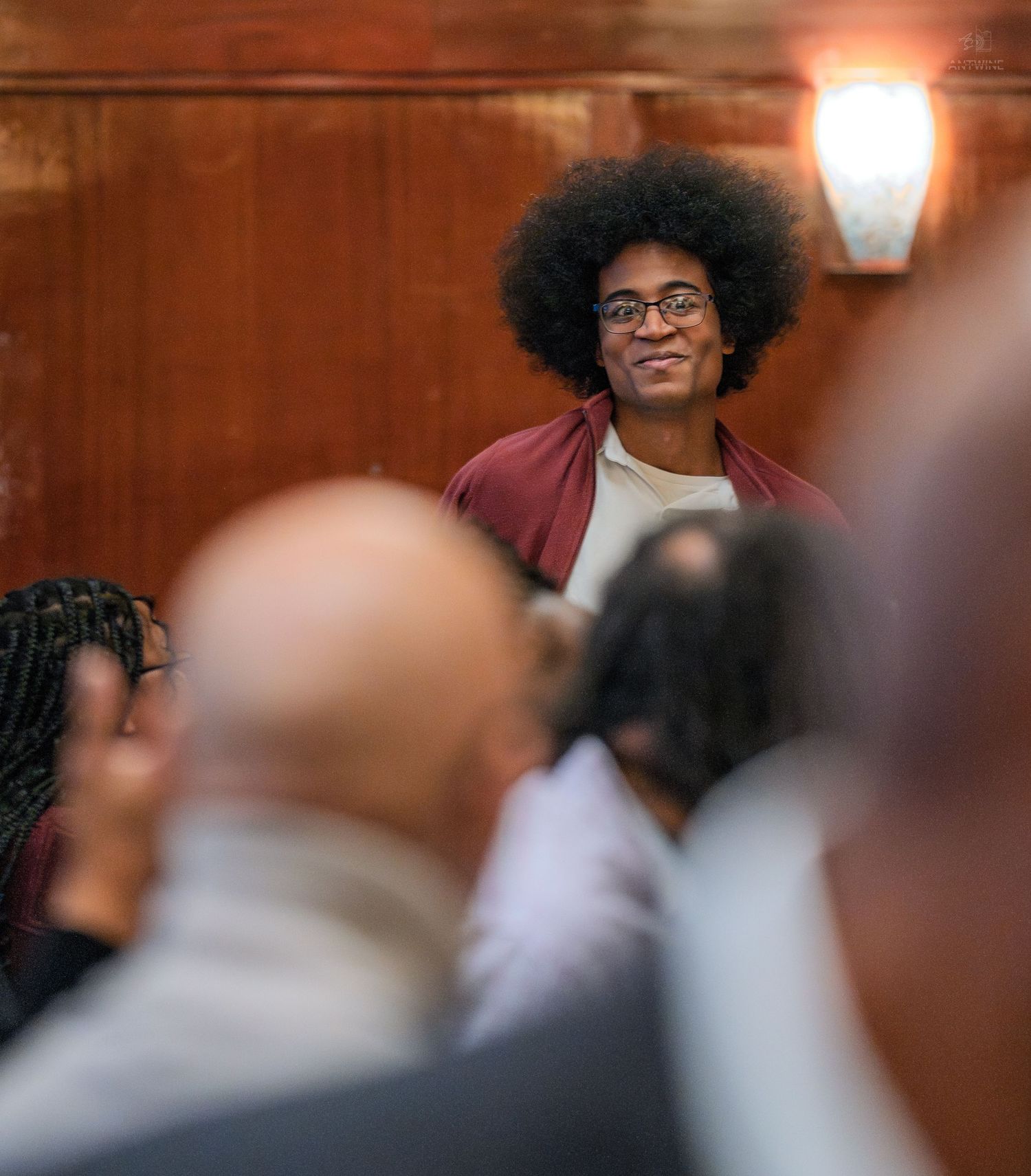 Smiling young person with afro and glasses stands out in a crowd at an indoor gathering.