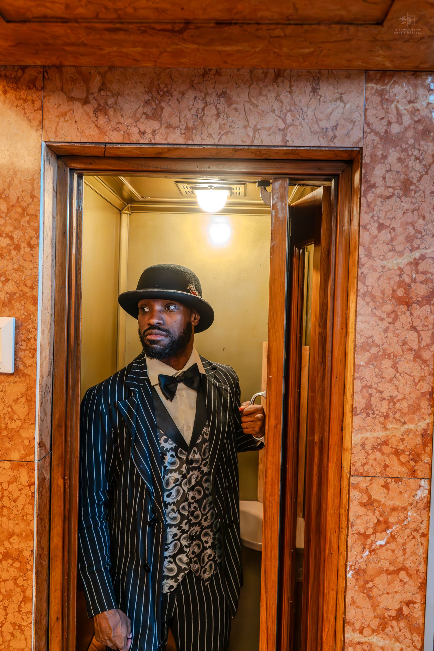 Stylish man in pinstripe suit, bow tie, and fedora hat standing in an elegant elevator with marble walls.