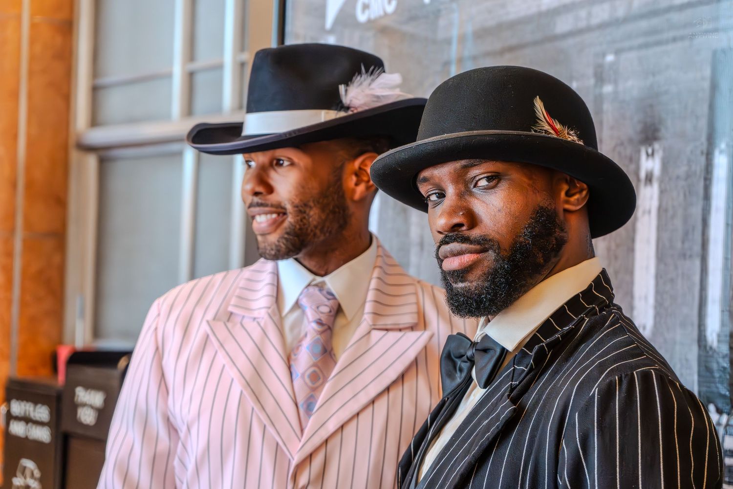 Two stylish men wearing wide-brim hats and pinstripe suits pose together at a formal event.